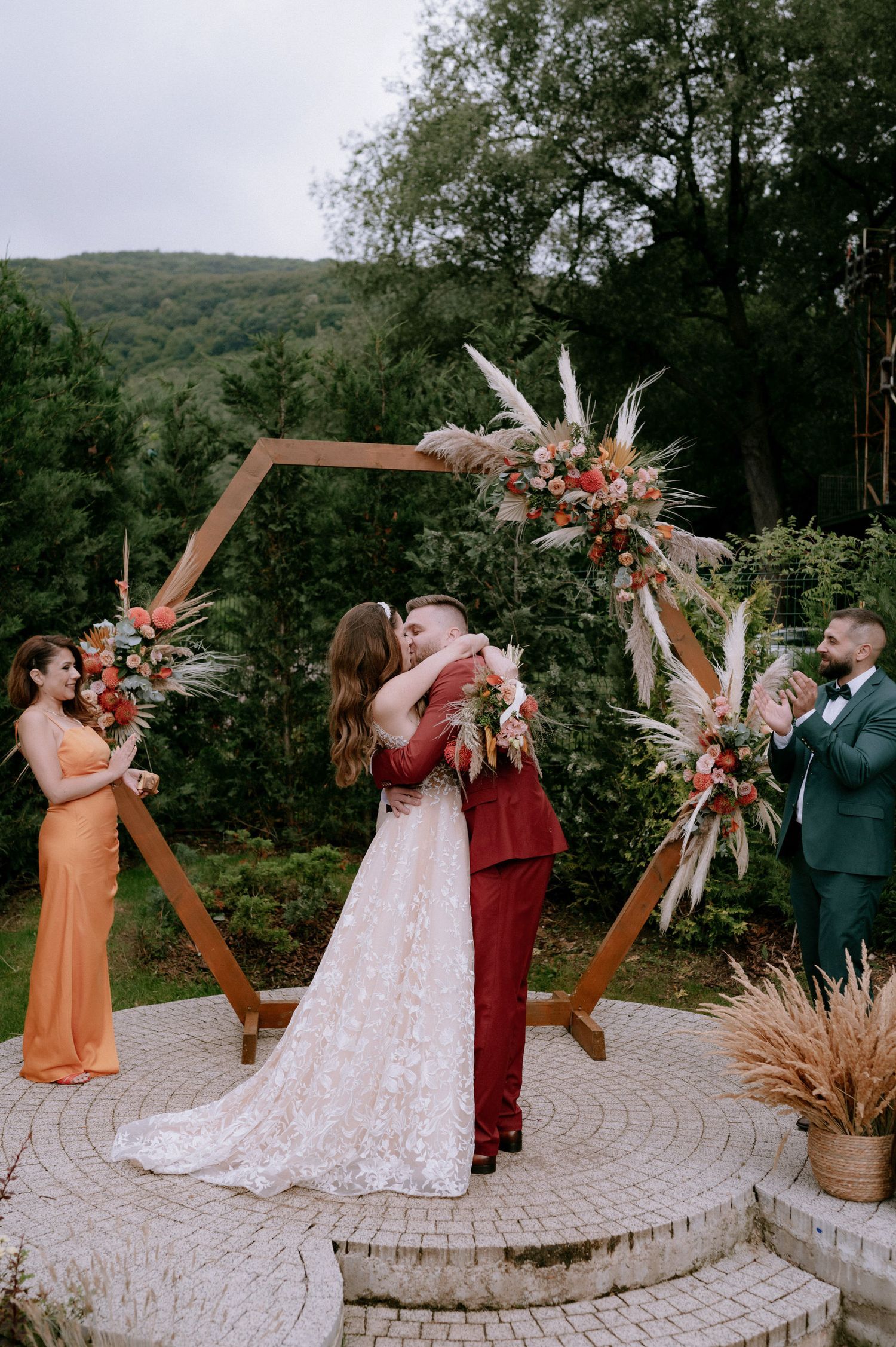 Wedding party members take celebratory shots together on a circular stone patio surrounded by greenery and floral decor.