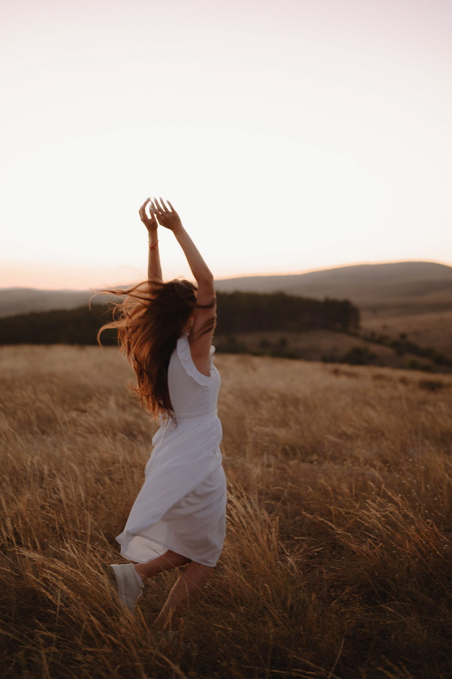 A person in a white dress dances in a golden field at sunset, with rolling hills in the background.
