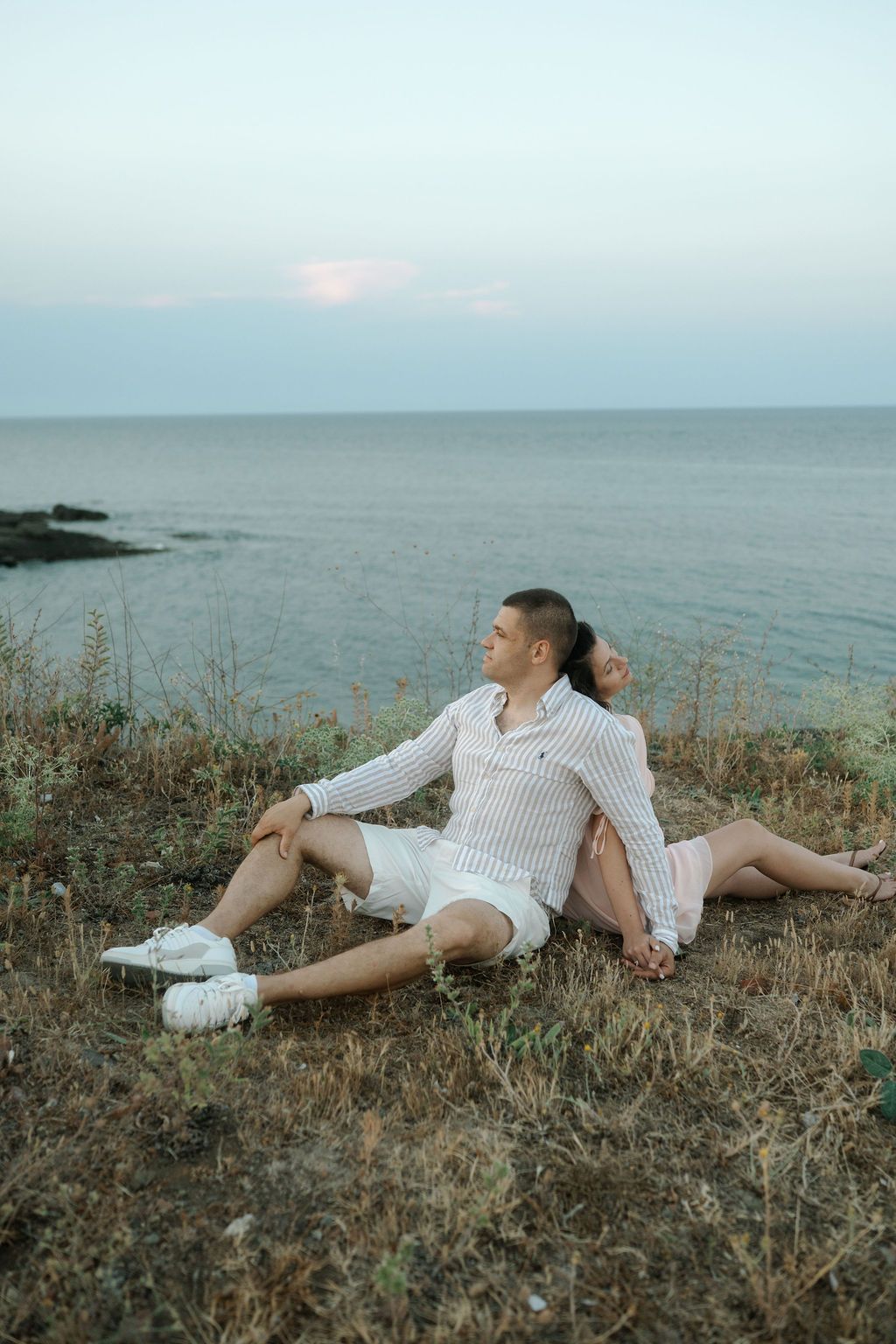 Someone wearing a white sweater lies on grassy coastal cliffs overlooking a misty ocean at sunset.