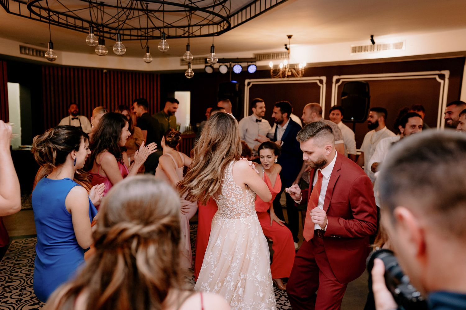 Wedding guests dancing and celebrating on a dimly lit dance floor at an indoor reception venue.