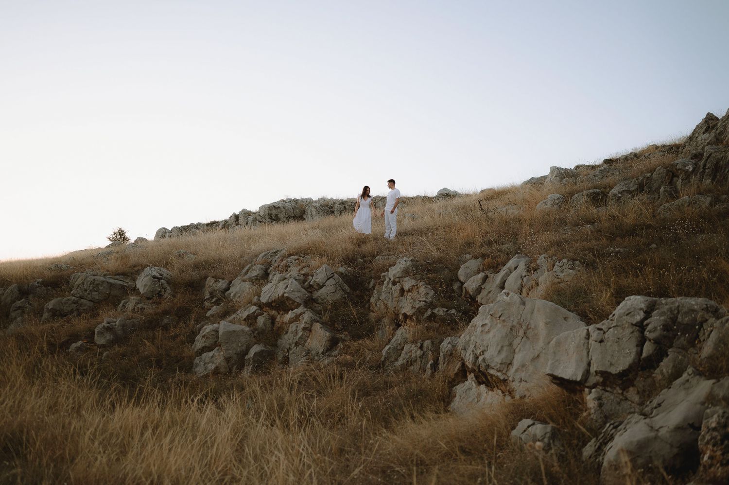 Two figures in white clothing stand on a rocky hillside covered in dry grass during golden hour.
