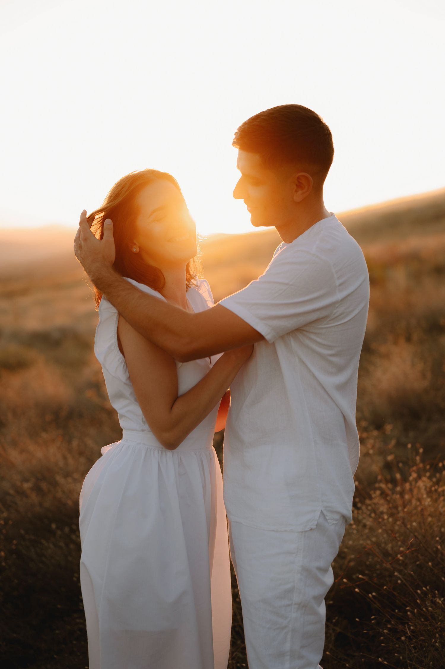 A couple in white outfits embrace during golden hour sunset in a field creating a romantic silhouette.