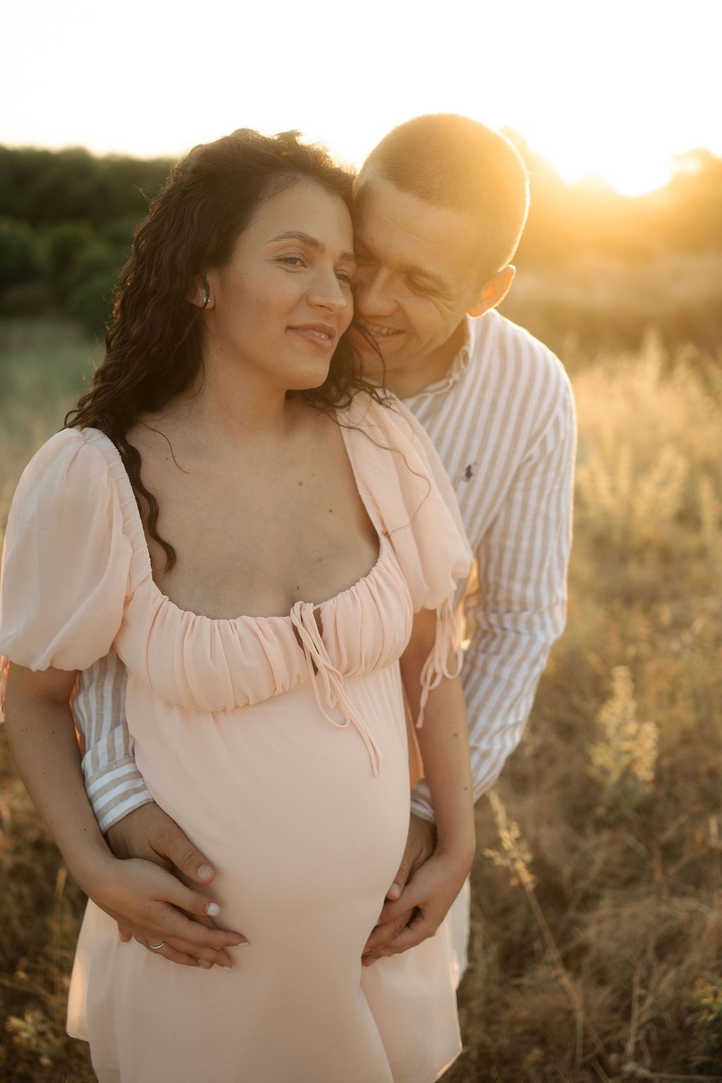 Couple embraces in golden sunset light while standing in a wildflower field wearing light colored clothing.