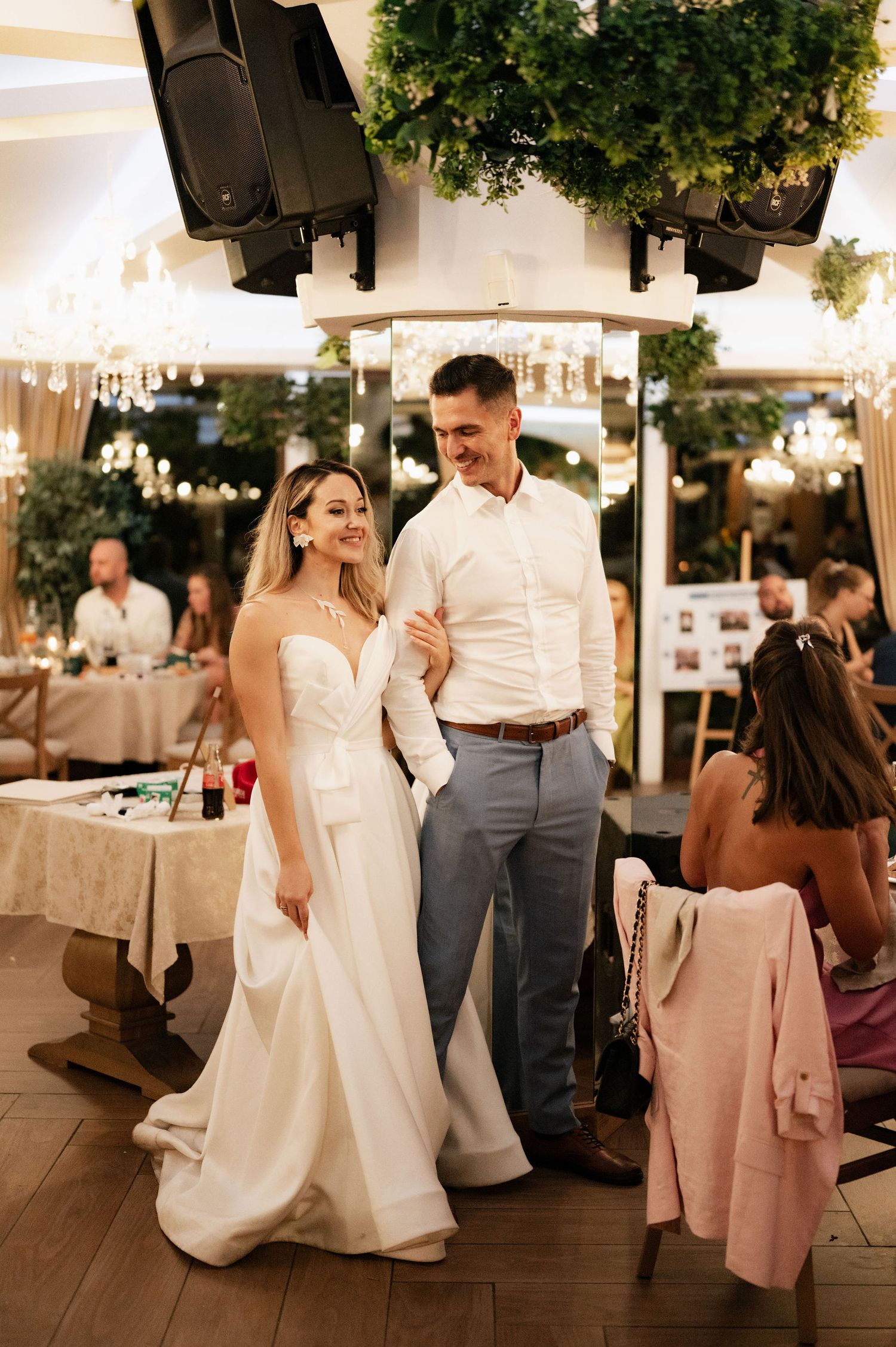 Wedding reception guests enjoying an evening celebration under white tent with greenery and string light decorations.