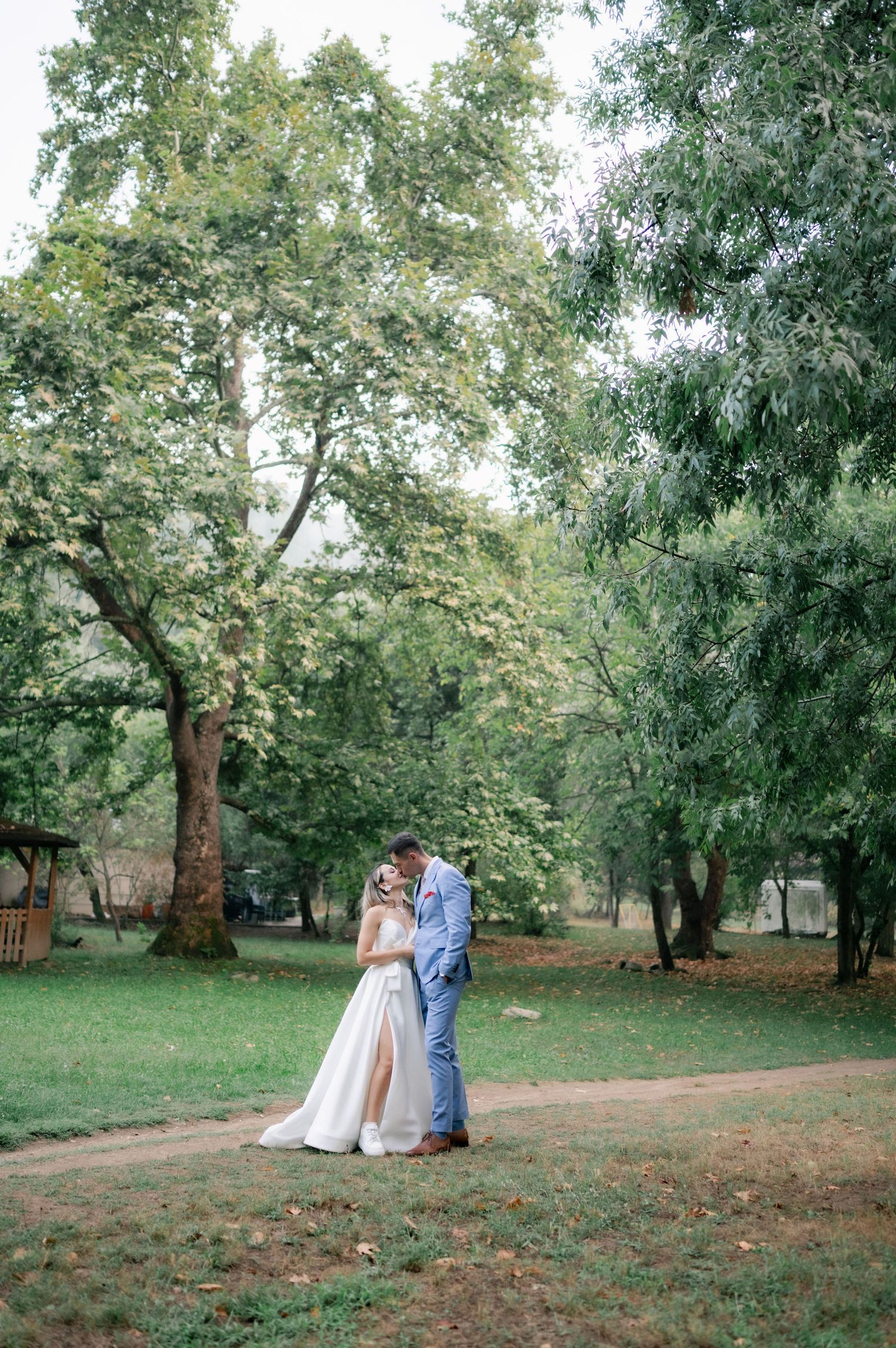 A couple in wedding attire strolls through a serene garden setting with tall trees and dappled sunlight.