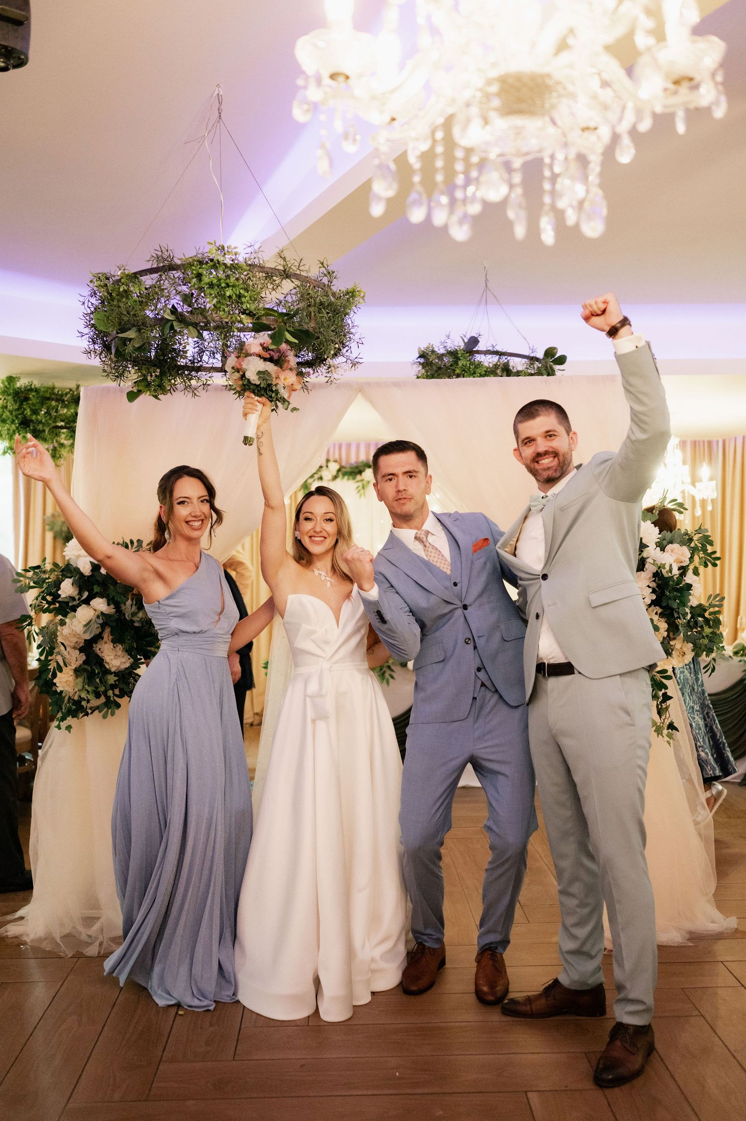 Wedding party in gray suits and lavender dresses celebrate joyfully with raised arms in an elegant venue.