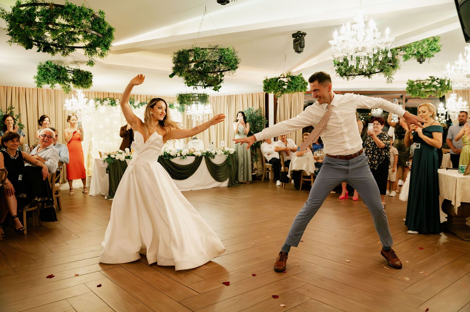 Wedding guests dance energetically in a reception hall decorated with hanging greenery and white decor.