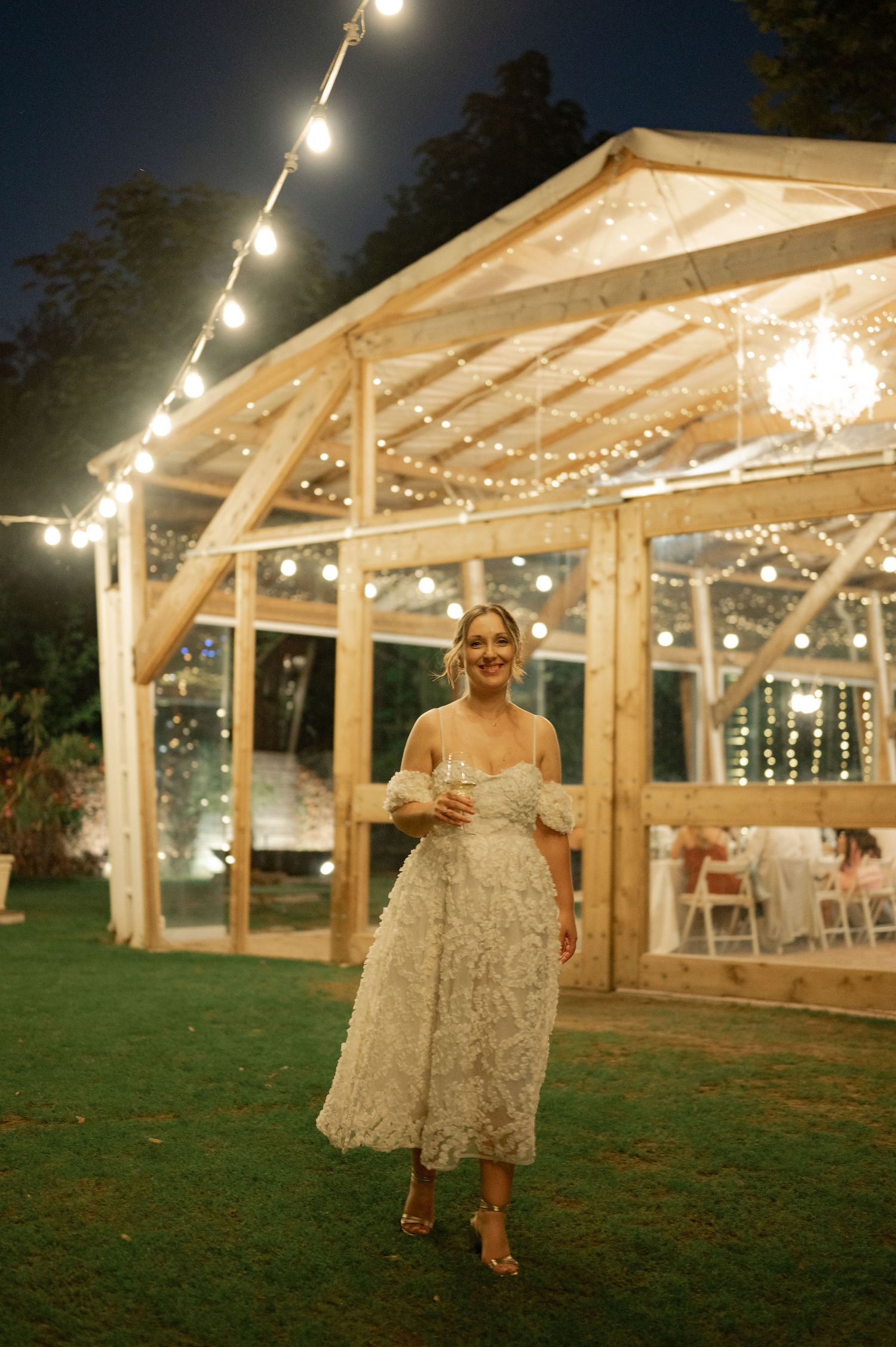 A lit-up outdoor wedding tent with string lights glows warmly against the evening sky.