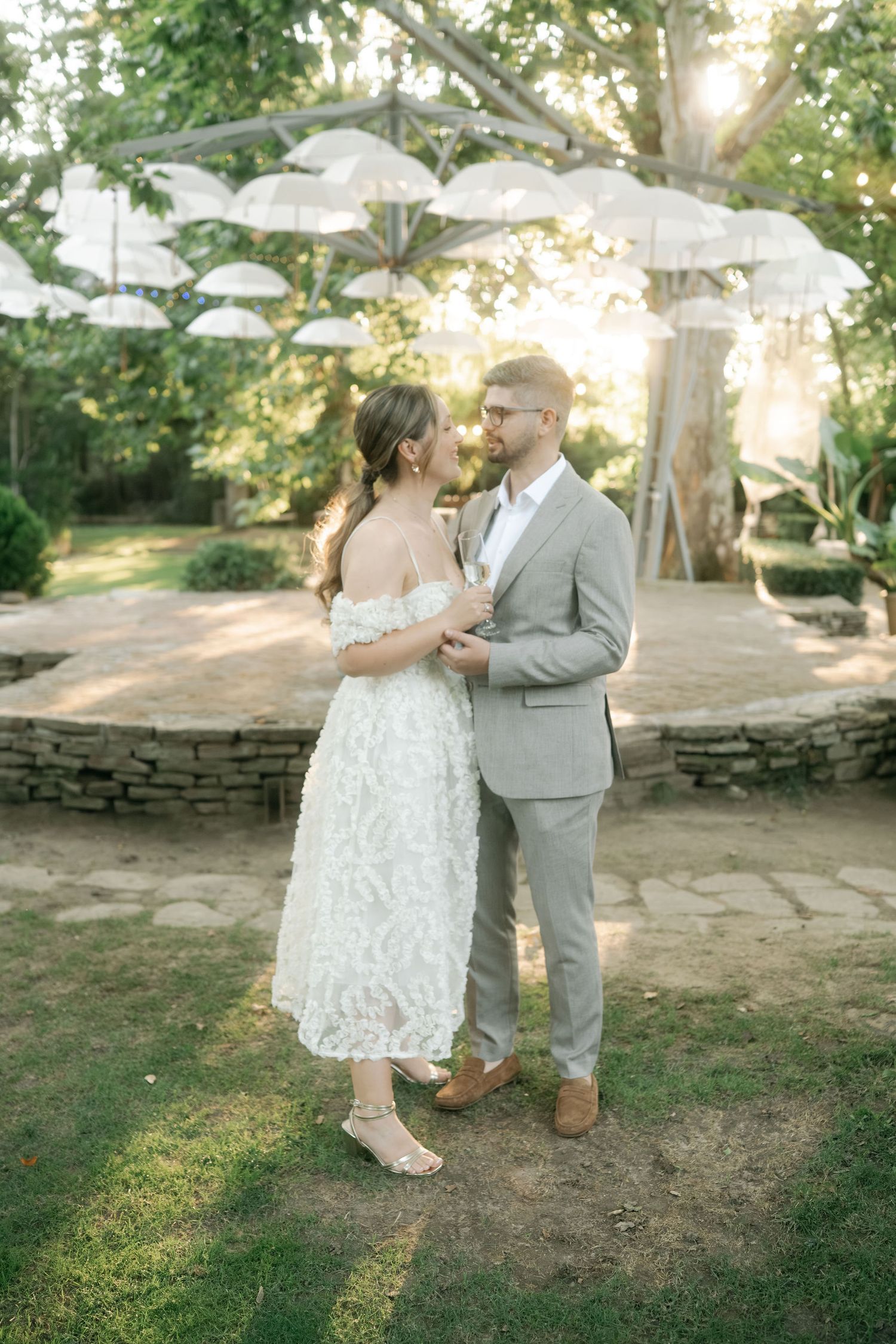 A romantic outdoor wedding scene with white umbrella decorations hanging overhead and sunlight filtering through trees.