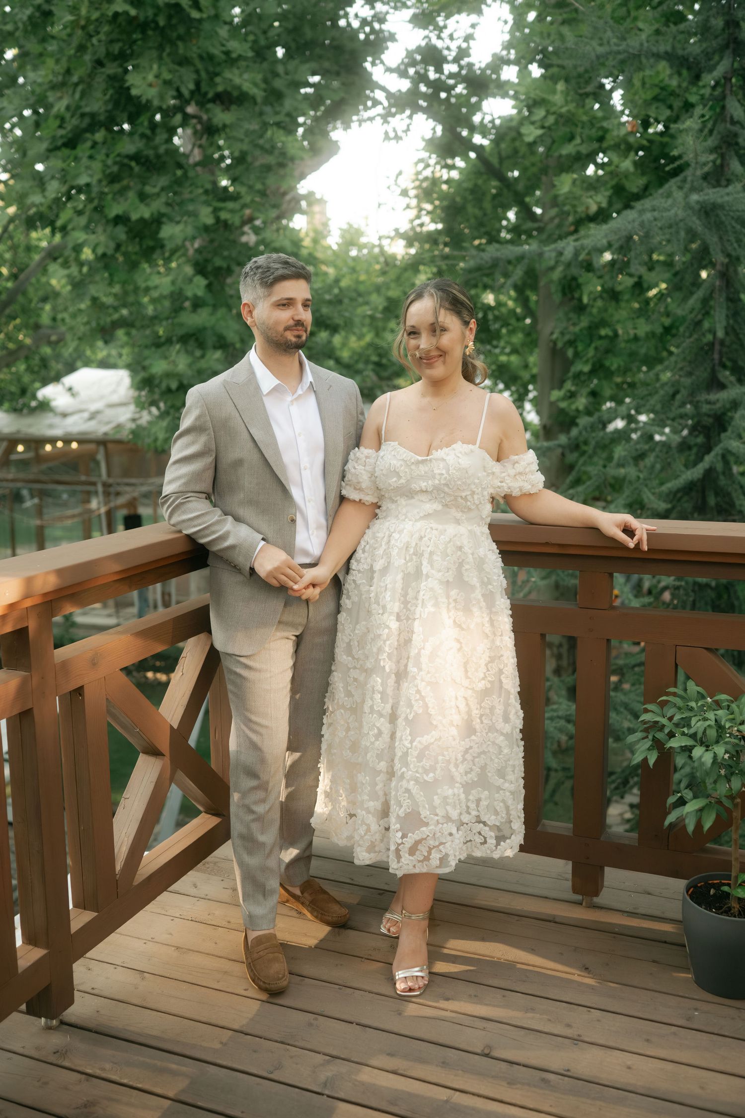 Romantic couple in formal attire shares playful moments on a wooden deck surrounded by lush greenery.