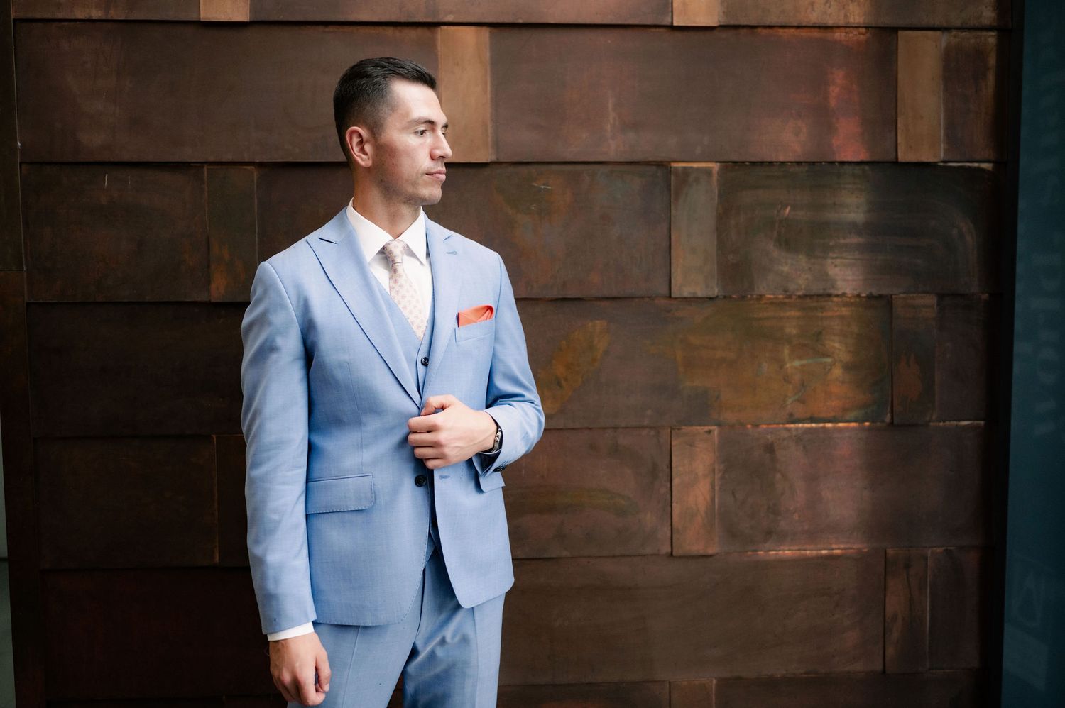 A person in a light blue suit and coral tie poses confidently against a dark wooden wall.