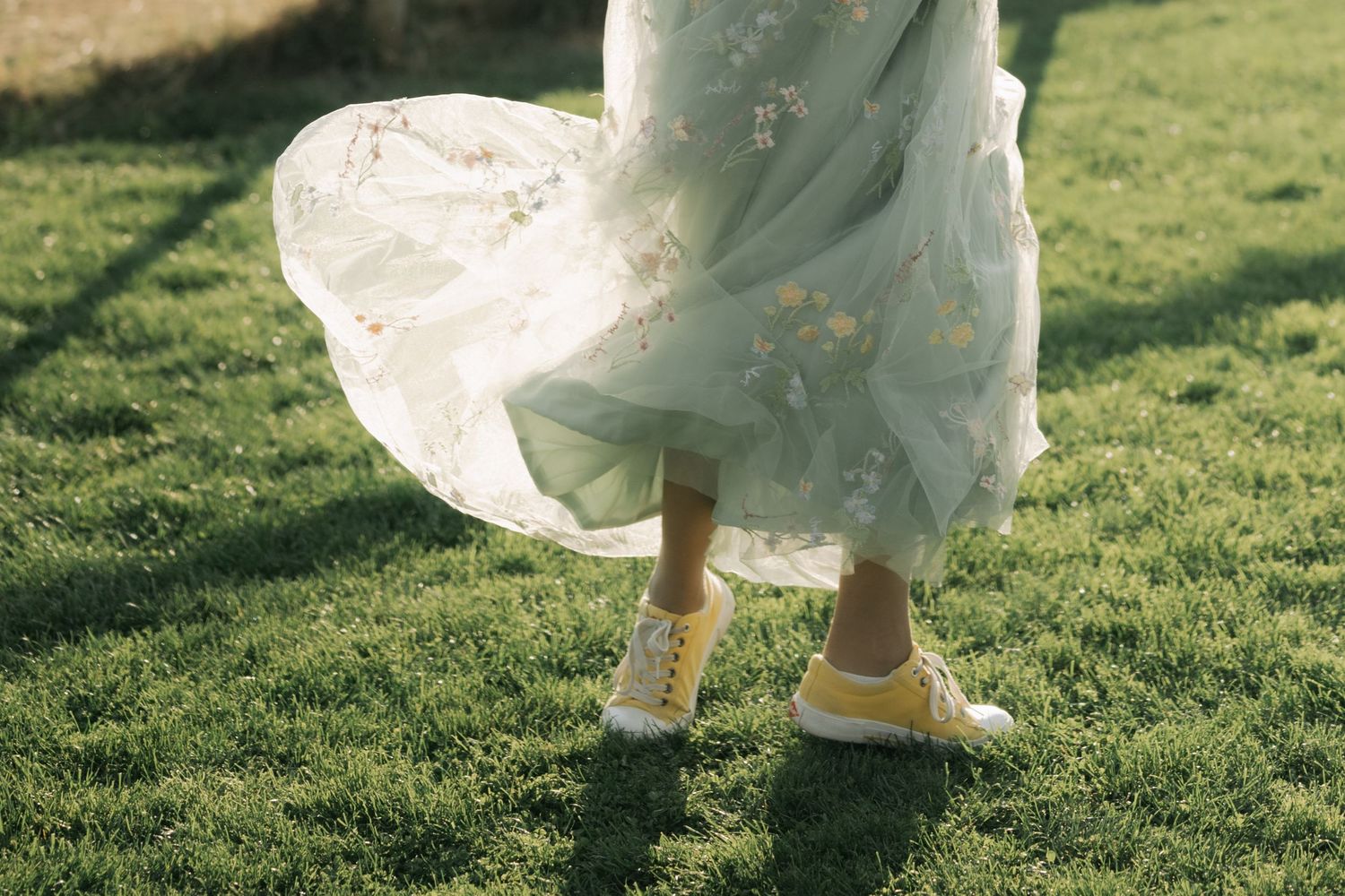 Close-up view of white wedding dress and gold heels walking on lush green grass in sunlight.