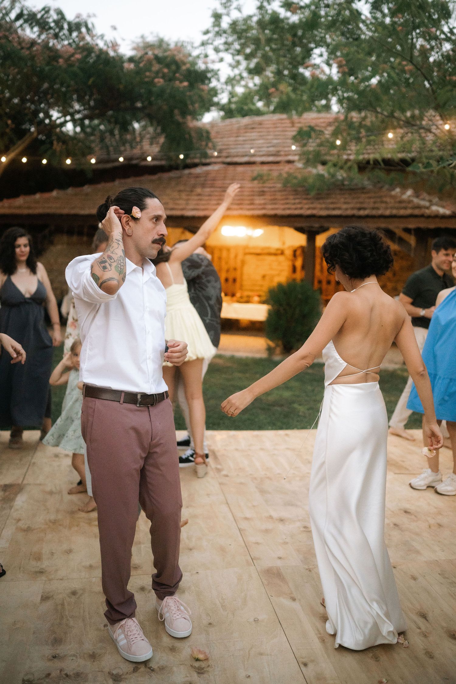 A couple dances joyfully together on a wooden deck at an outdoor wedding reception with string lights overhead.