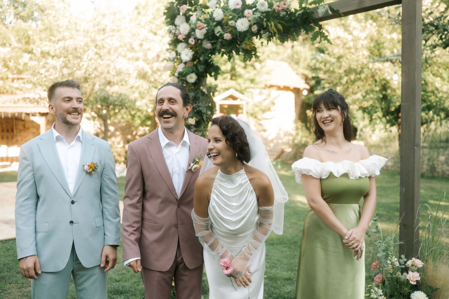 Wedding party members laugh and smile together while posing outdoors in formal attire during a sunny afternoon celebration.