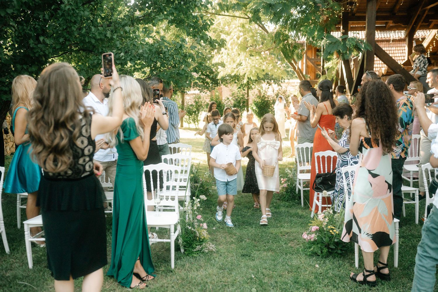 Wedding guests mingle during an outdoor garden reception with white chairs and emerald green bridesmaid dresses.