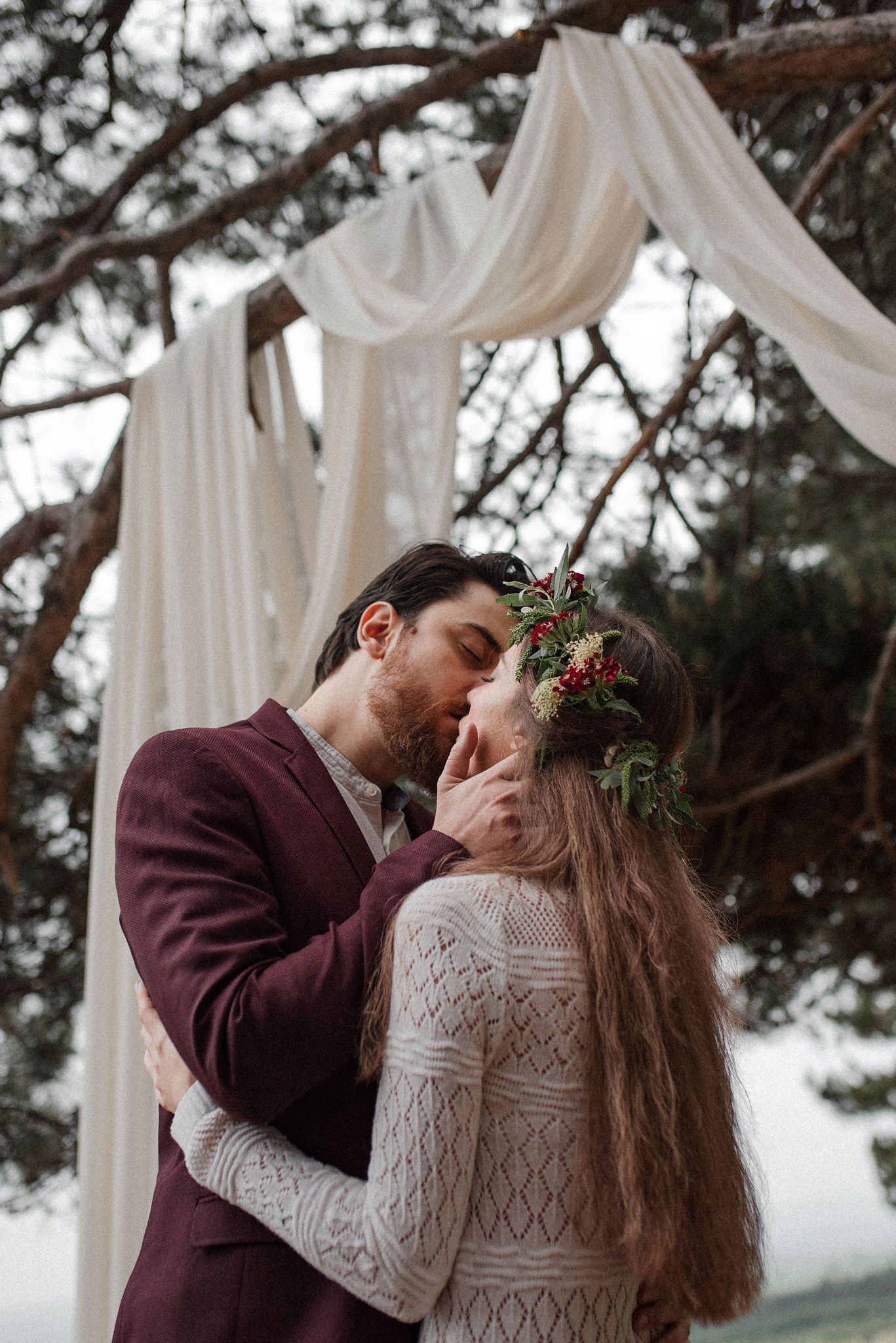 Romantic winter wedding couple embraces under white fabric draped between trees in an outdoor setting.
