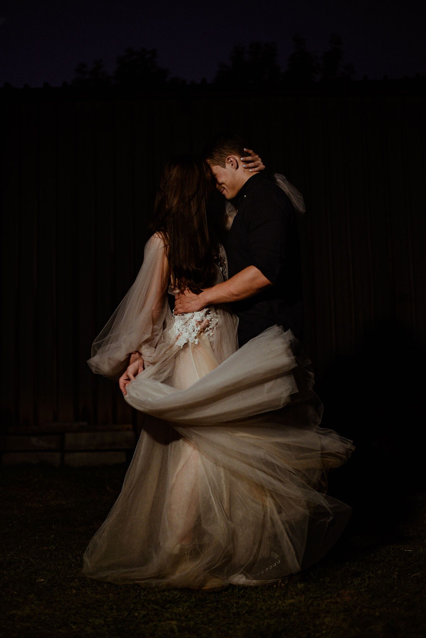 A romantic wedding couple dances together in a dimly lit outdoor setting with flowing white dress movements.