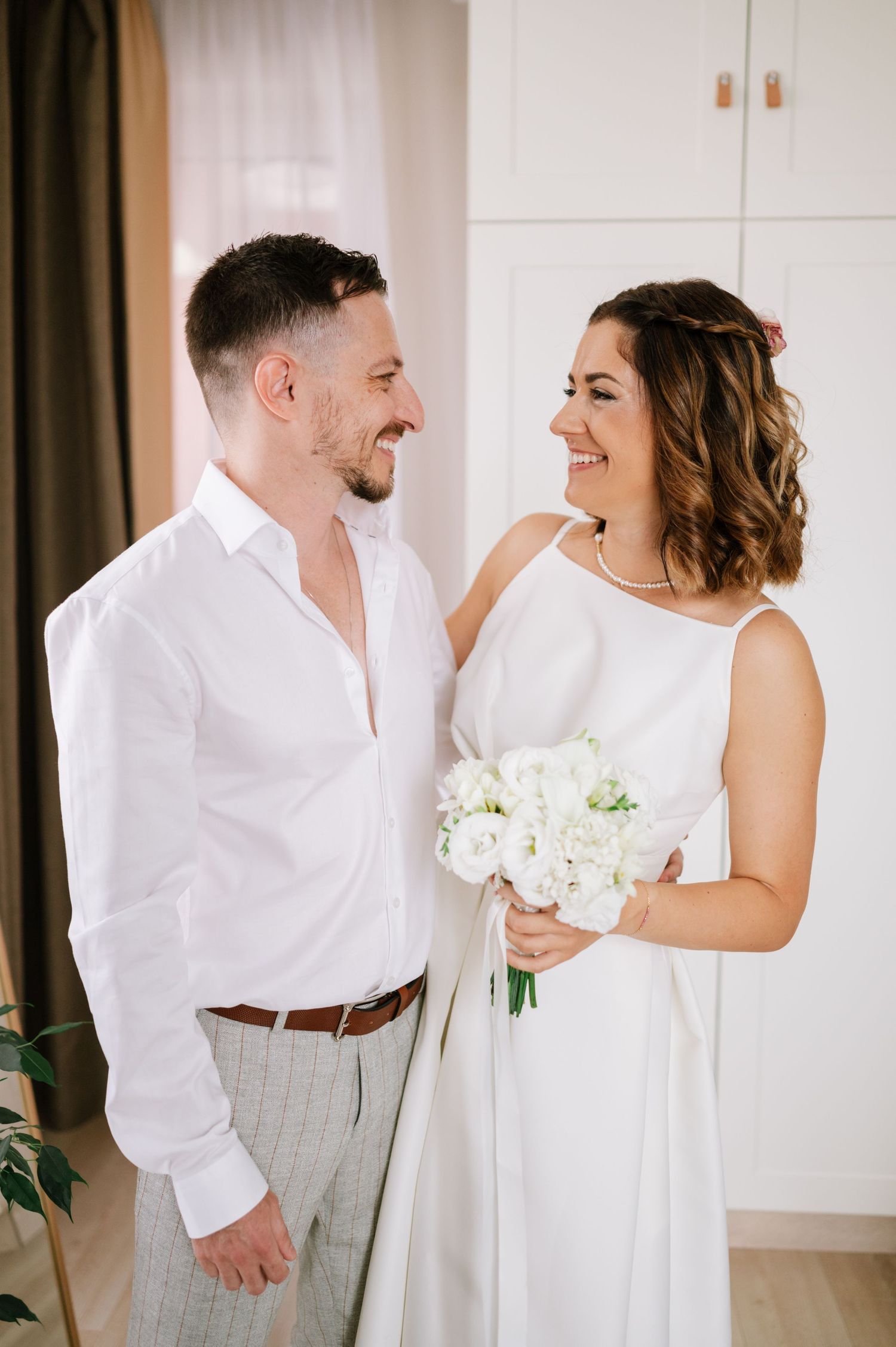 A couple shares joyful moments in white attire during an indoor photo session with elegant curtains and decor.
