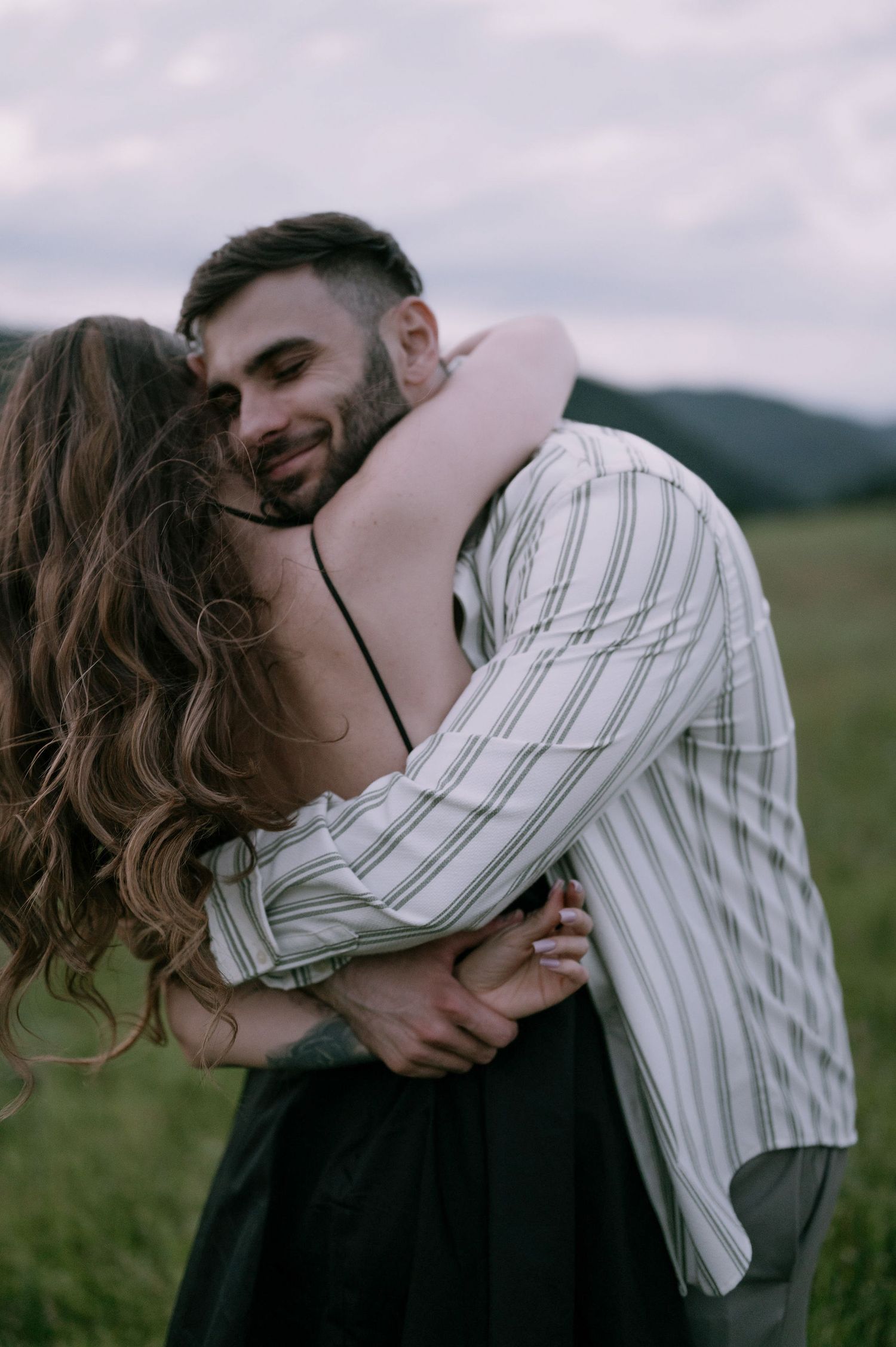 A couple embraces intimately in a field with rolling hills in the background on a cloudy day.