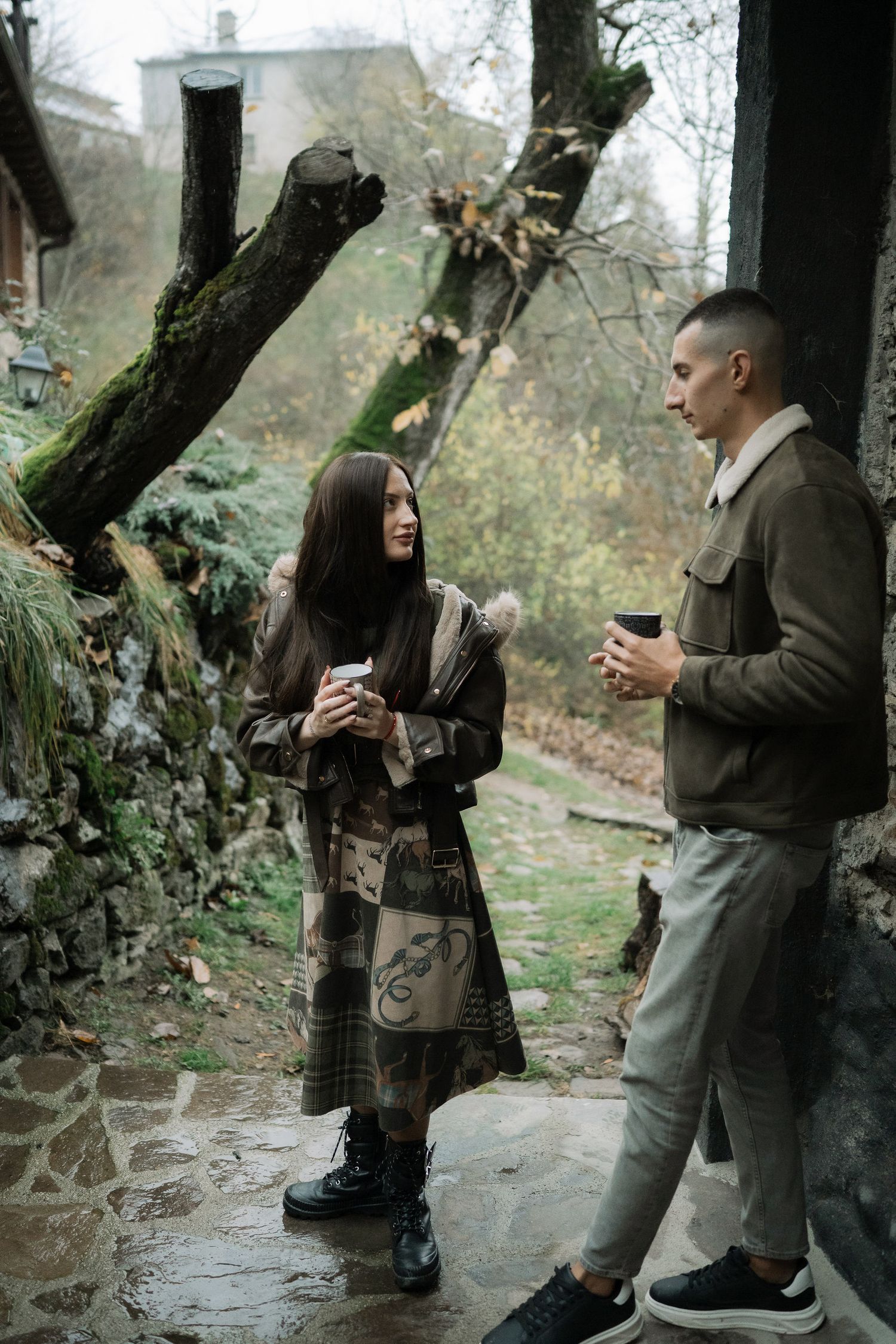 Two people stand together under trees on a stone path while holding drinks and having a conversation.