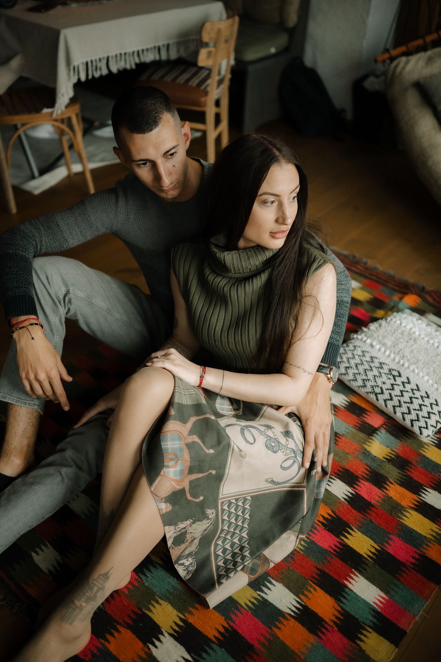 A couple sits on a colorful patterned rug in a dimly lit room with a sleeveless turtleneck and casual attire.