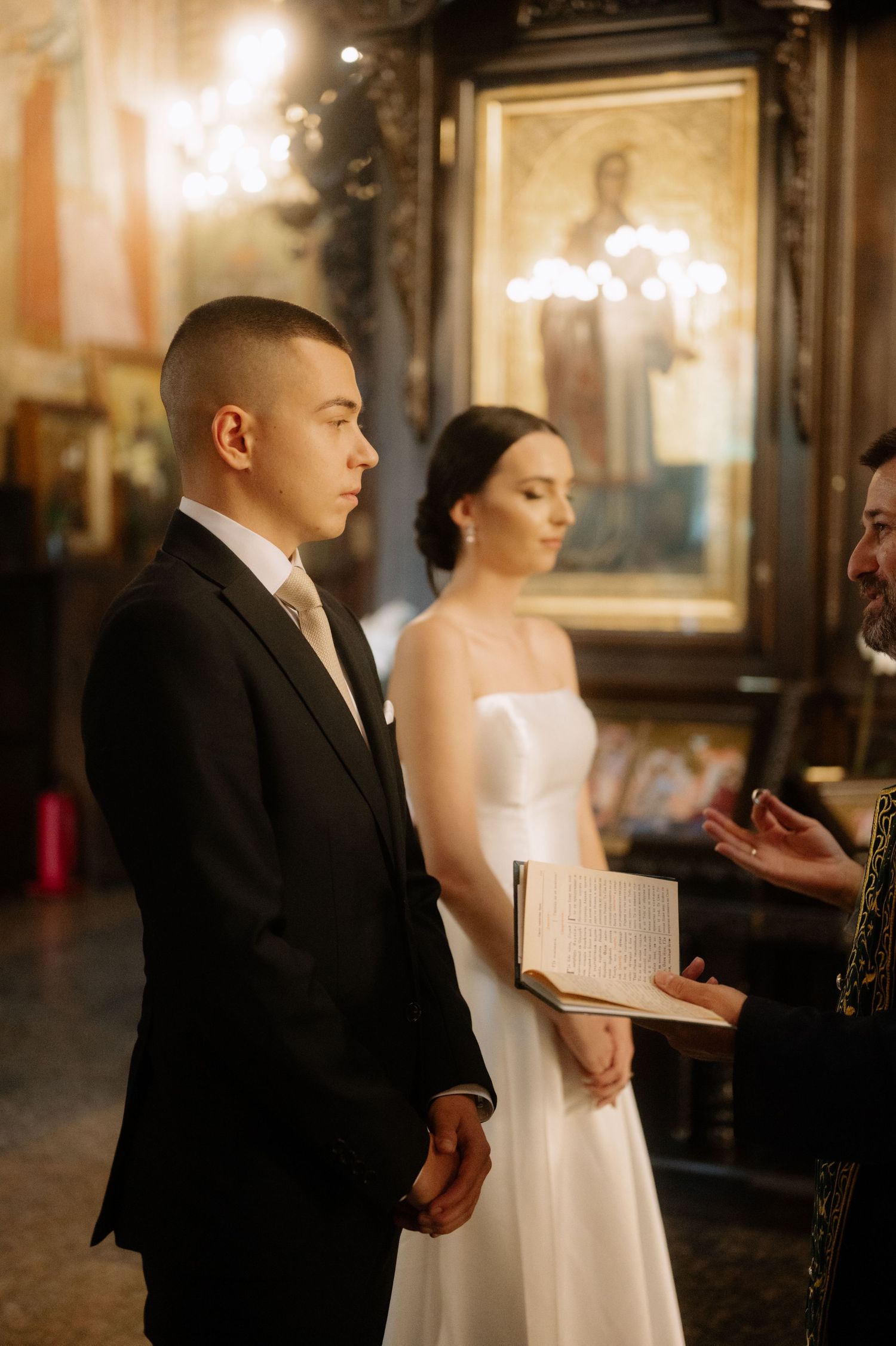 A bride and groom stand side by side during their wedding ceremony in an elegant indoor venue with warm lighting.