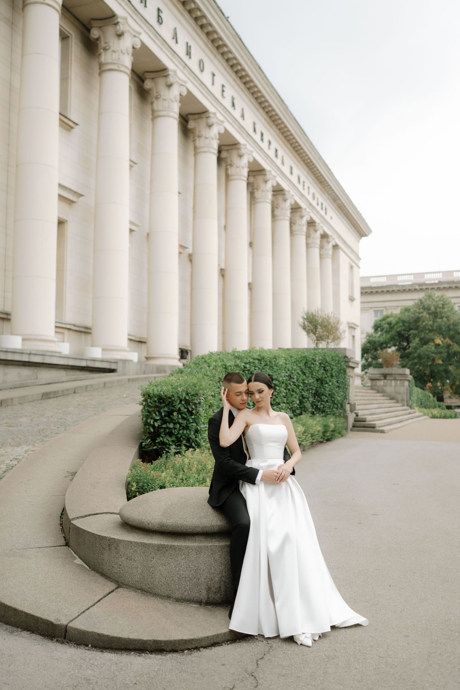 A couple poses outside a grand neoclassical building with white columns and manicured hedges.