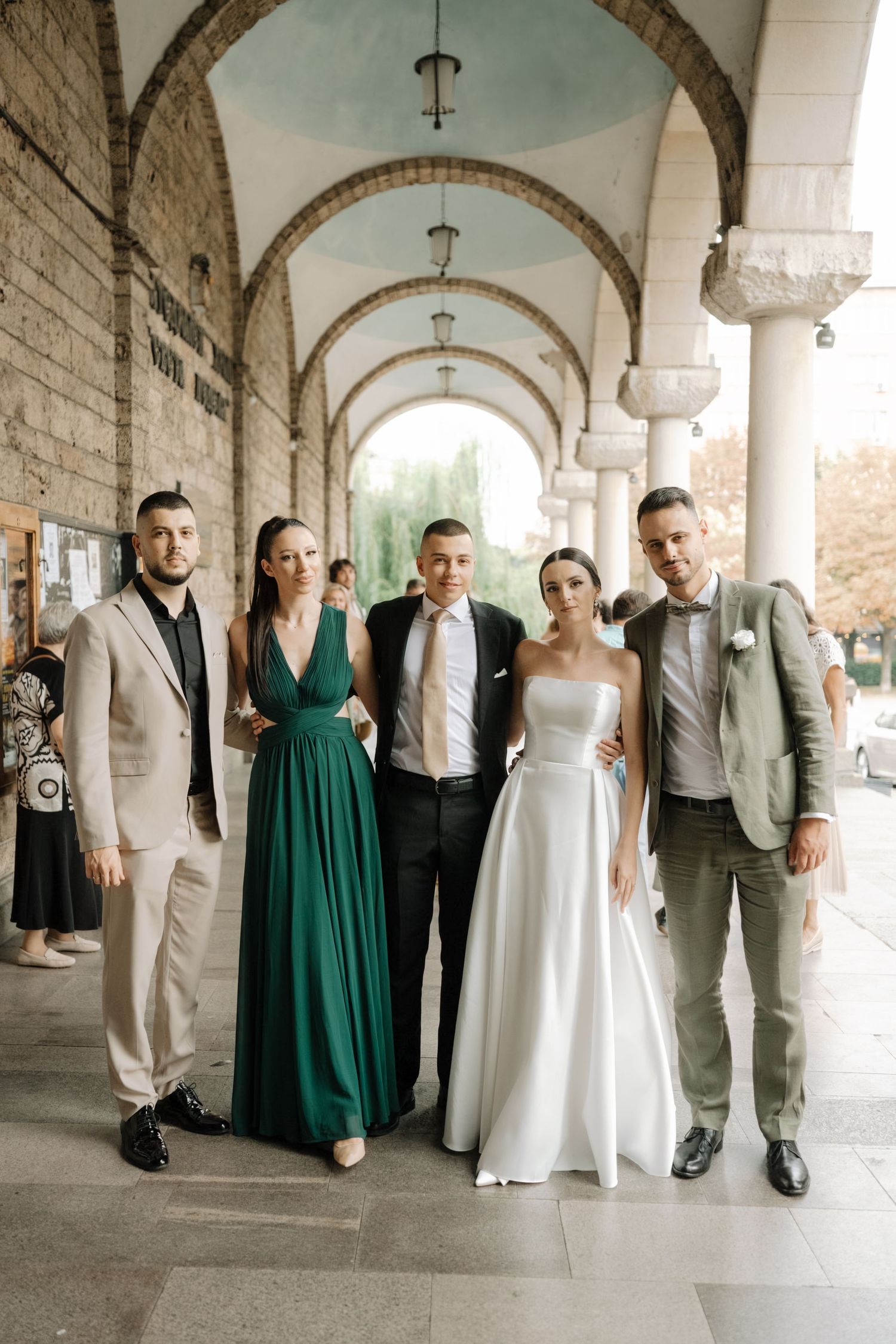 Wedding party in formal attire poses under arched colonnade with stone columns and vaulted ceiling.