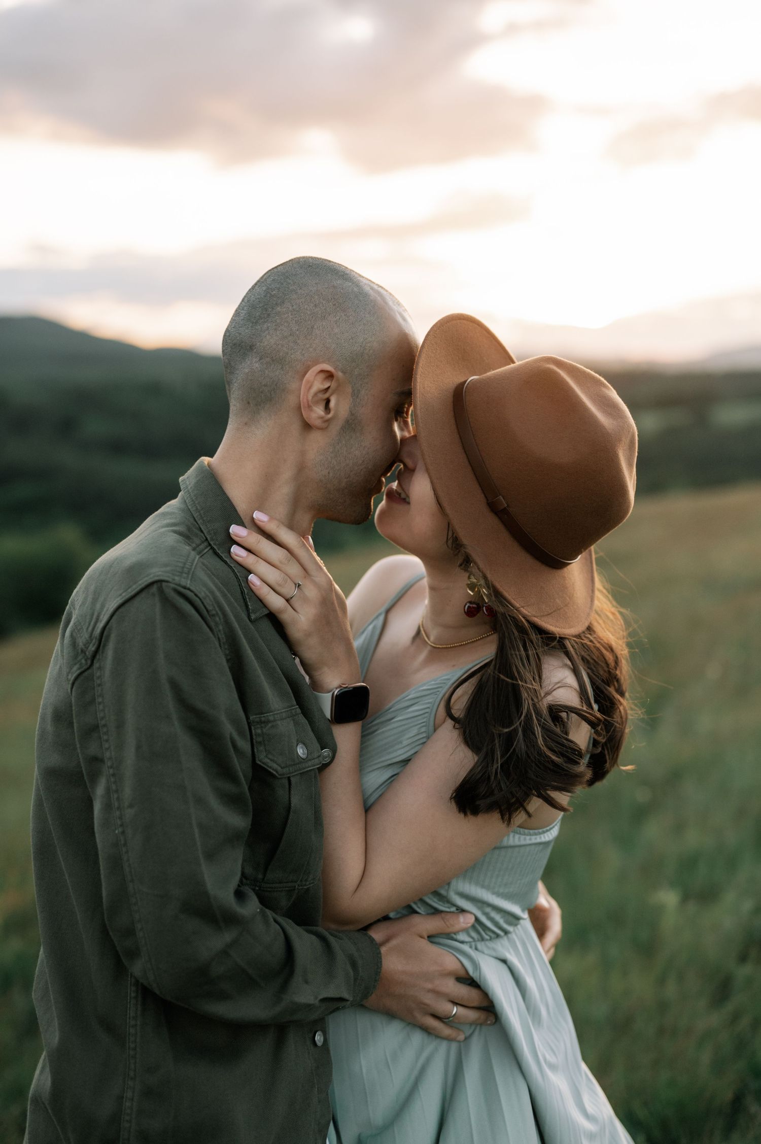 Couple sharing a romantic kiss at sunset in a green field, woman wearing a brown hat and light blue dress.