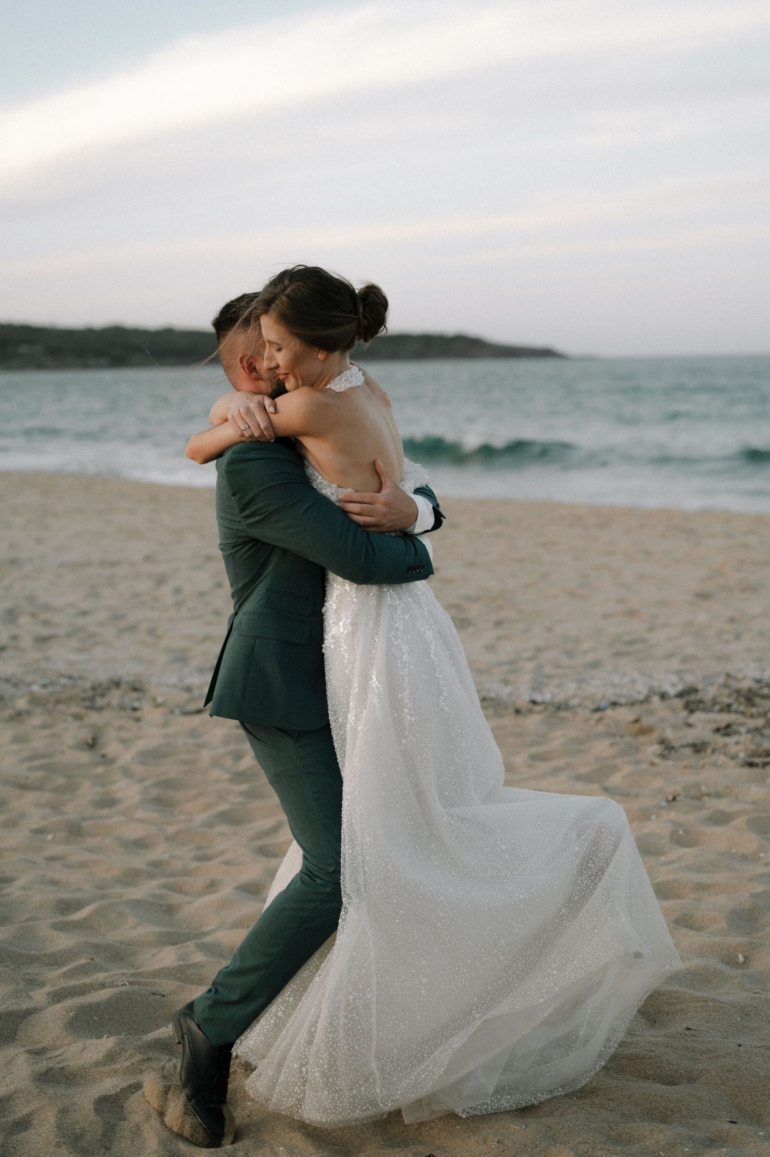 Bride and groom embracing on a sandy beach at sunset, bride in white gown, groom in green suit.