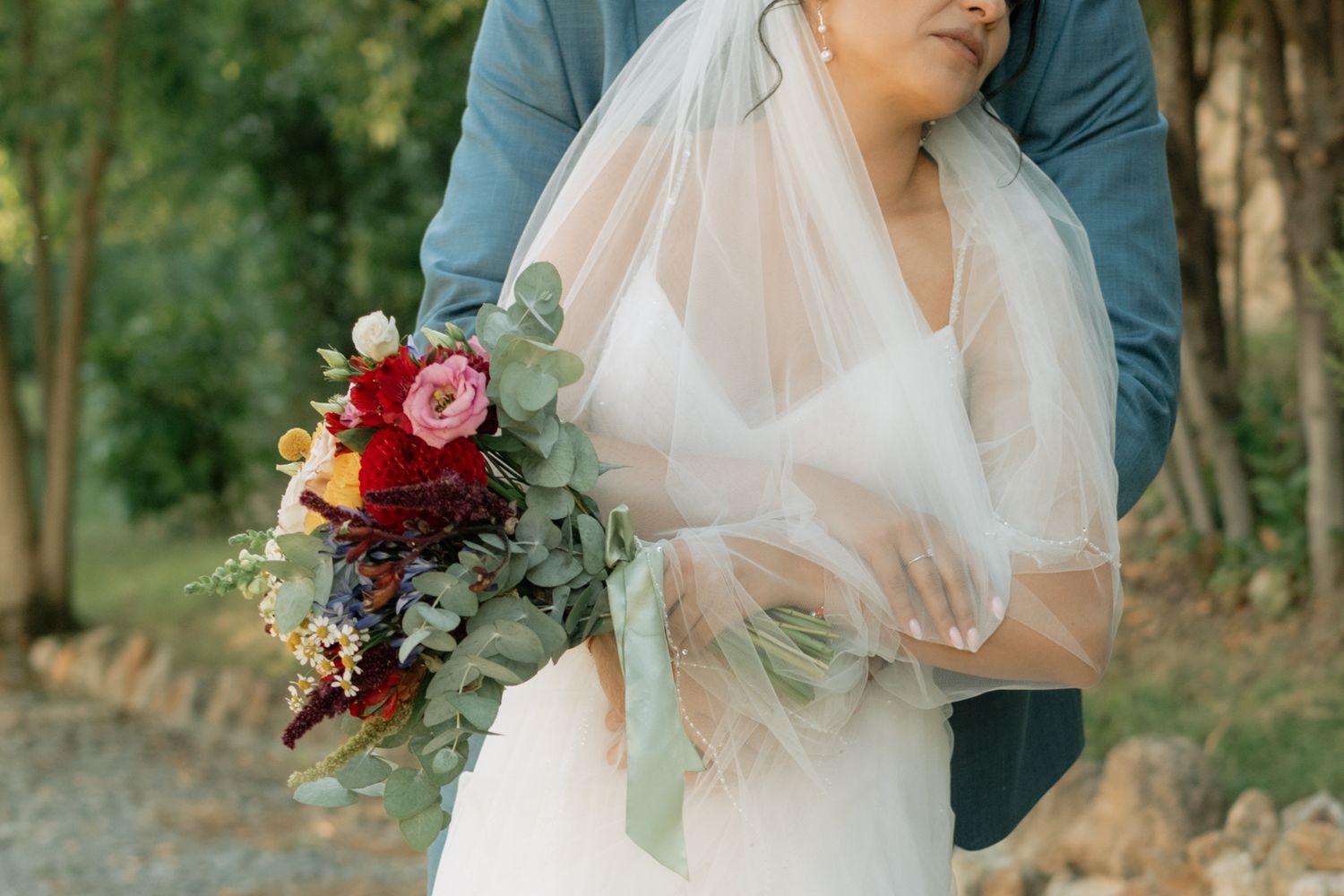 A couple in wedding attire embraces in a lush forest setting with soft natural lighting and floral bouquet details.