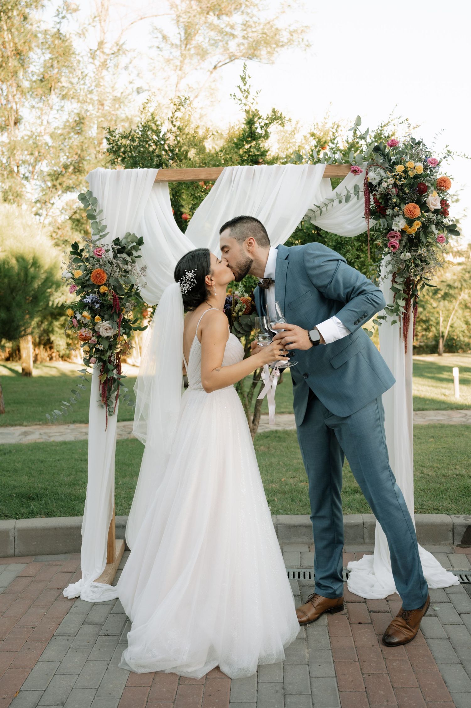 A couple shares a playful moment in front of a white draped wedding arch decorated with coral flowers in an outdoor setting.