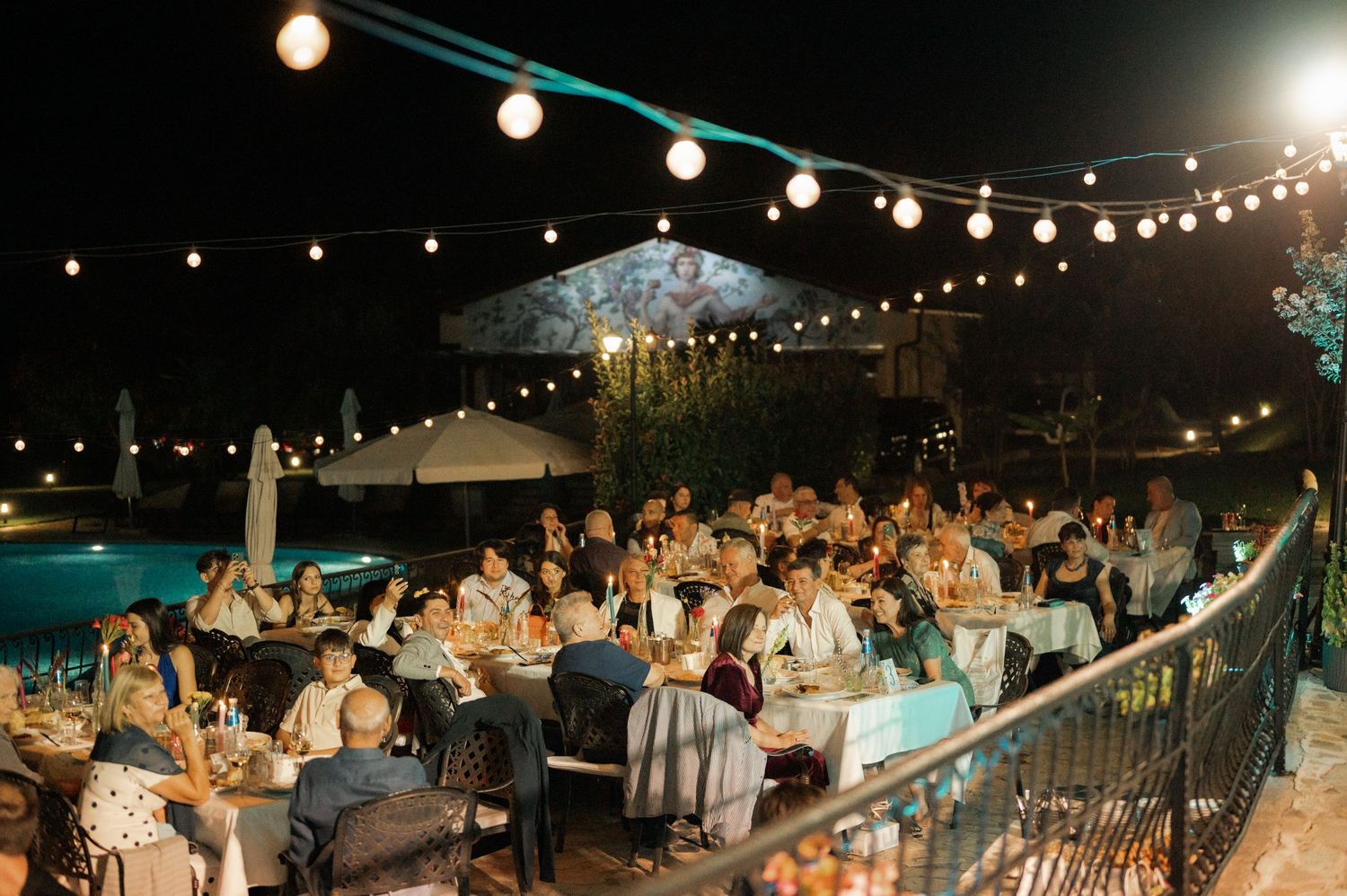 Evening outdoor event with string lights illuminating guests seated at tables beside a swimming pool.