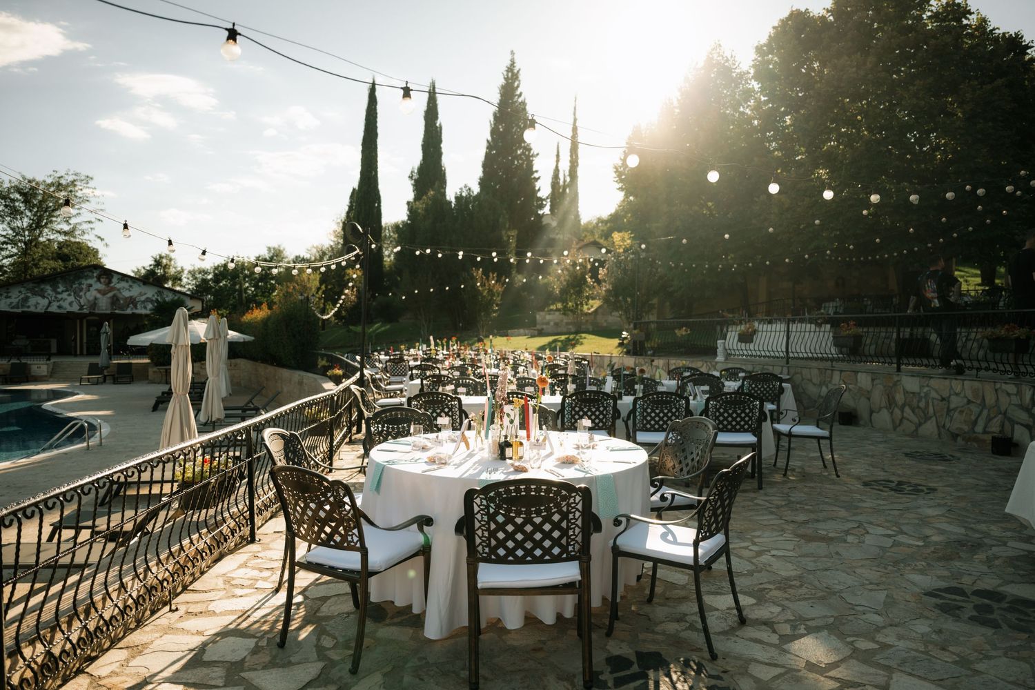 Outdoor wedding reception setup with string lights and tables on a stone patio at sunset with cypress trees in background.