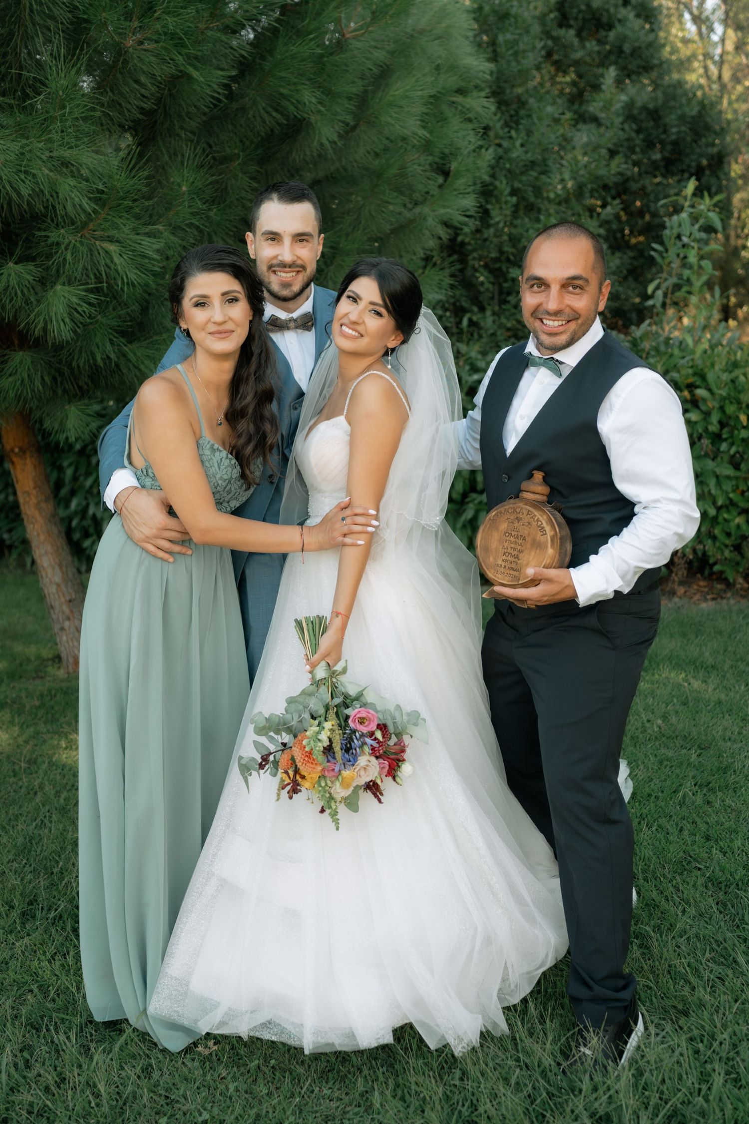 Wedding party poses together outdoors with bride in white gown holding colorful bouquet against green foliage backdrop.