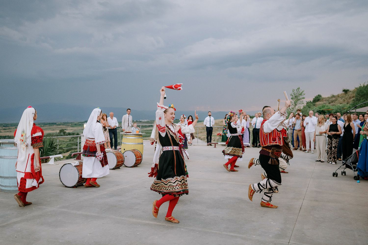 Folk dancers in traditional red and white costumes perform on an outdoor stage.
