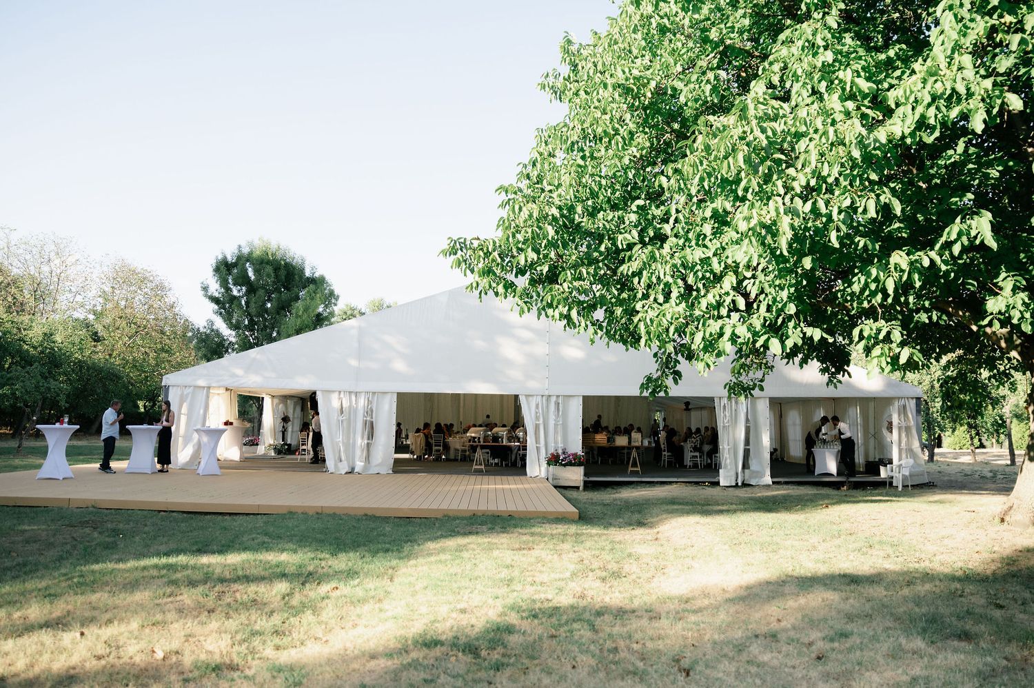 Large white wedding tent pavilion arranged in a grassy field surrounded by mature trees.