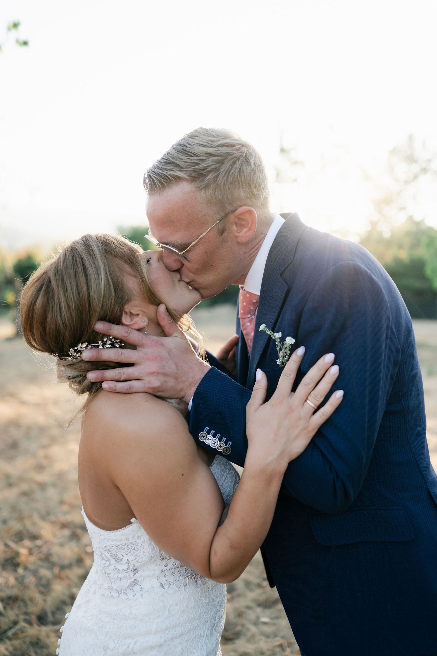 A tender kiss between newlyweds captured in warm sunlight among olive trees.