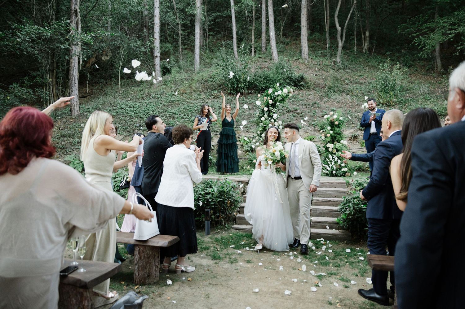 Wedding celebration in a wooded setting with guests gathered on rustic benches.