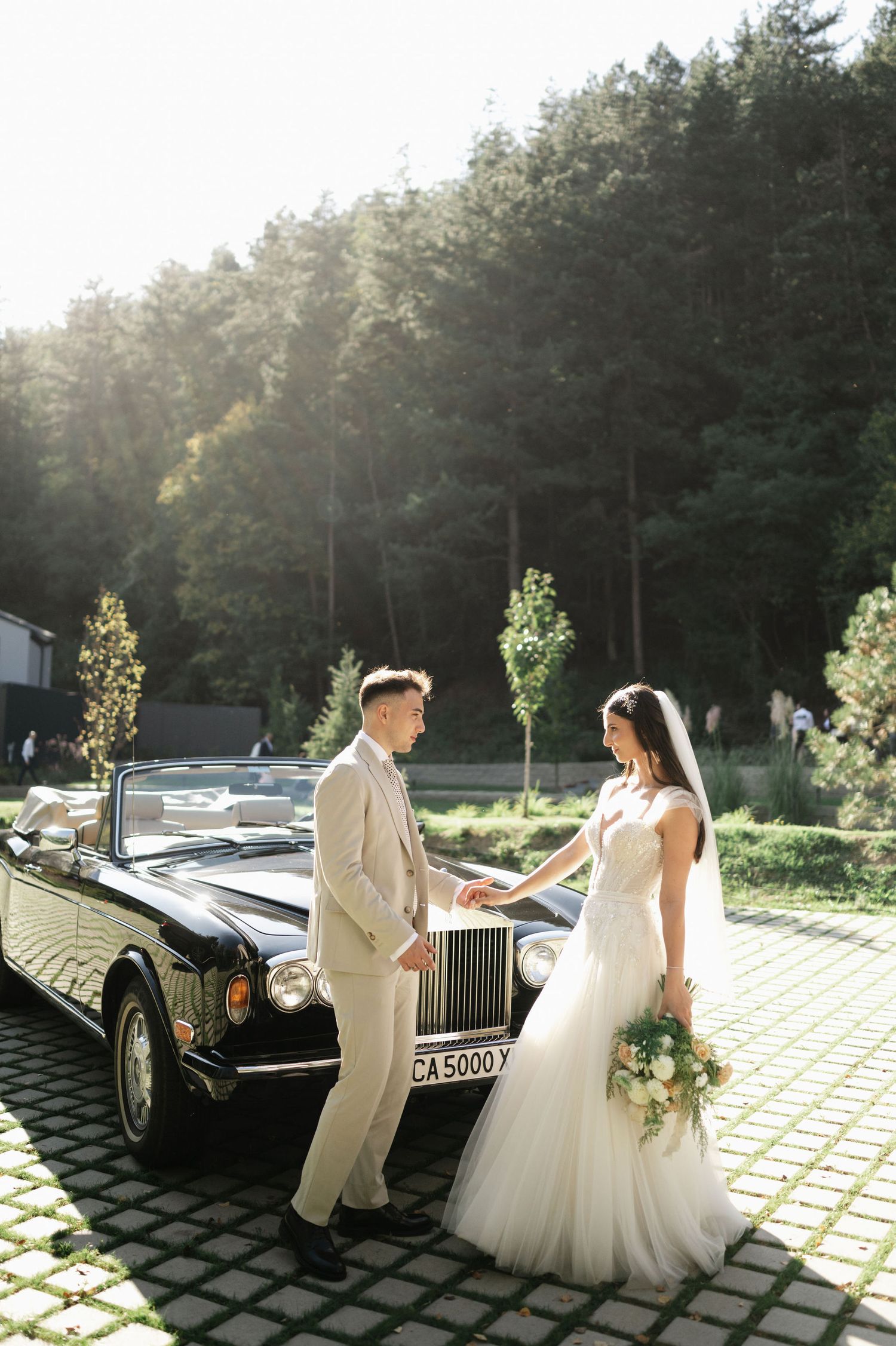 A wedding couple poses beside a classic black Rolls Royce convertible in a sunlit forest setting.