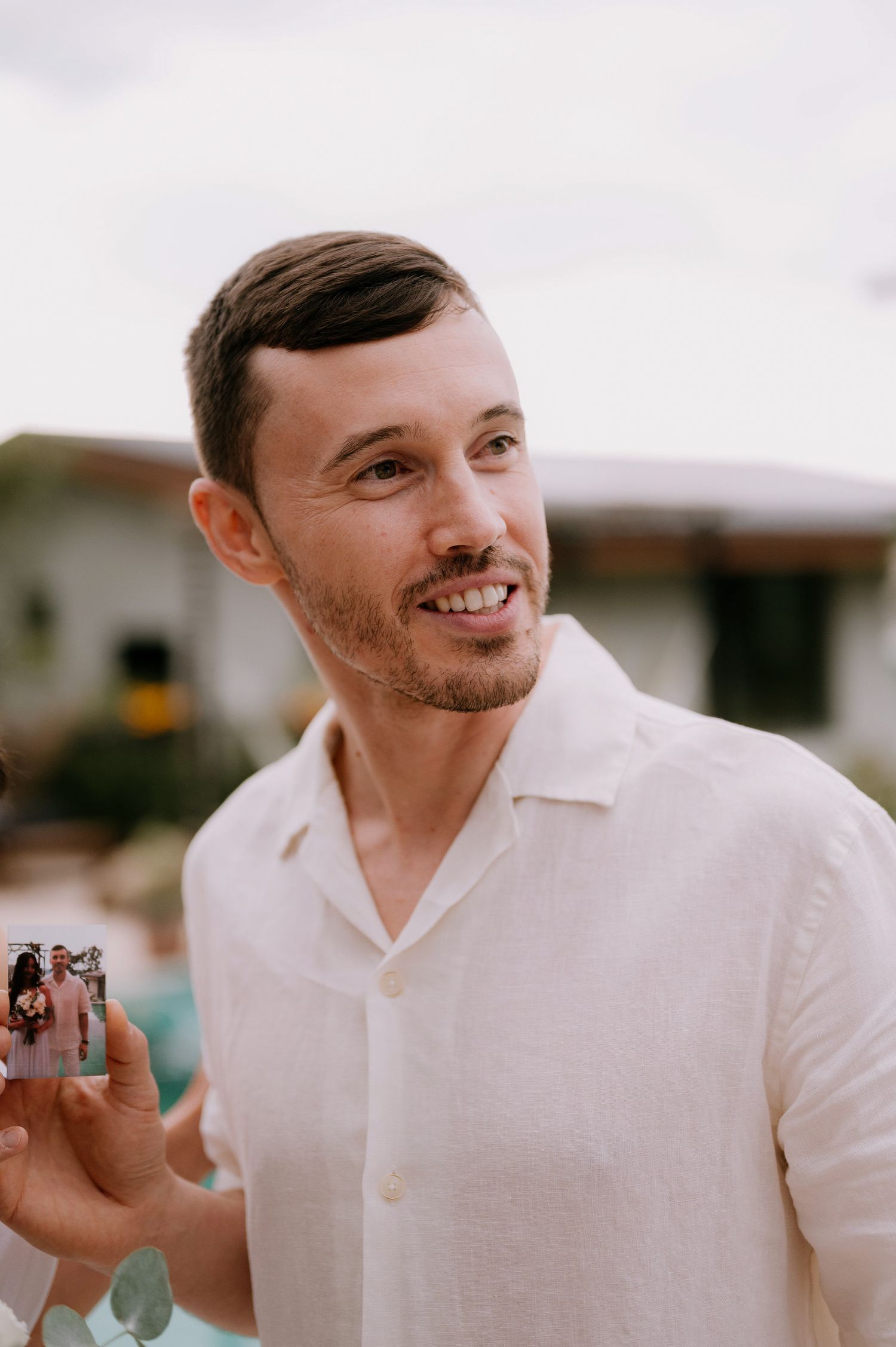 A person in a white shirt smiling while holding a small object outdoors.