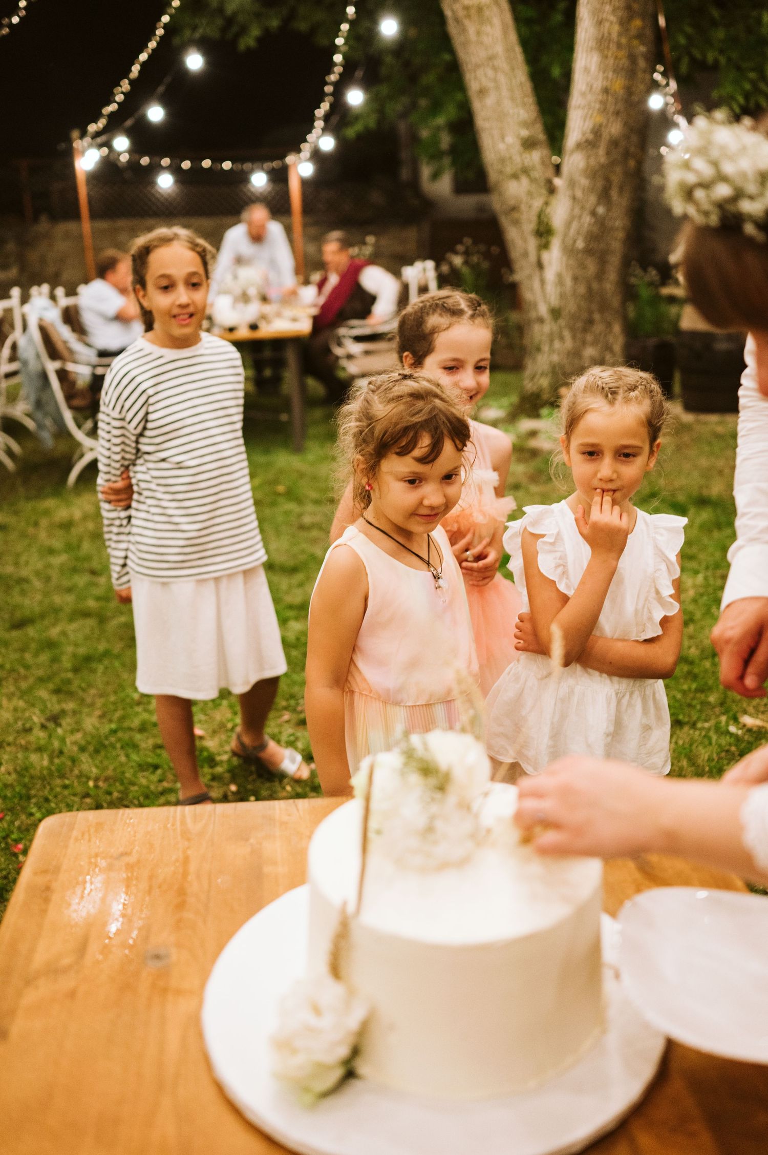 Children gather excitedly around a white wedding cake at an outdoor reception with string lights.