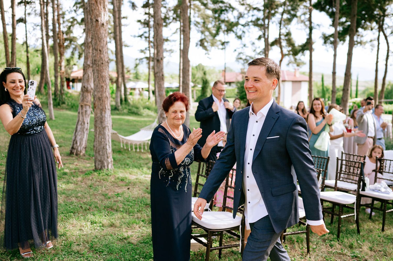 Wedding party celebrates in a wooded outdoor setting with dark chairs arranged for ceremony.