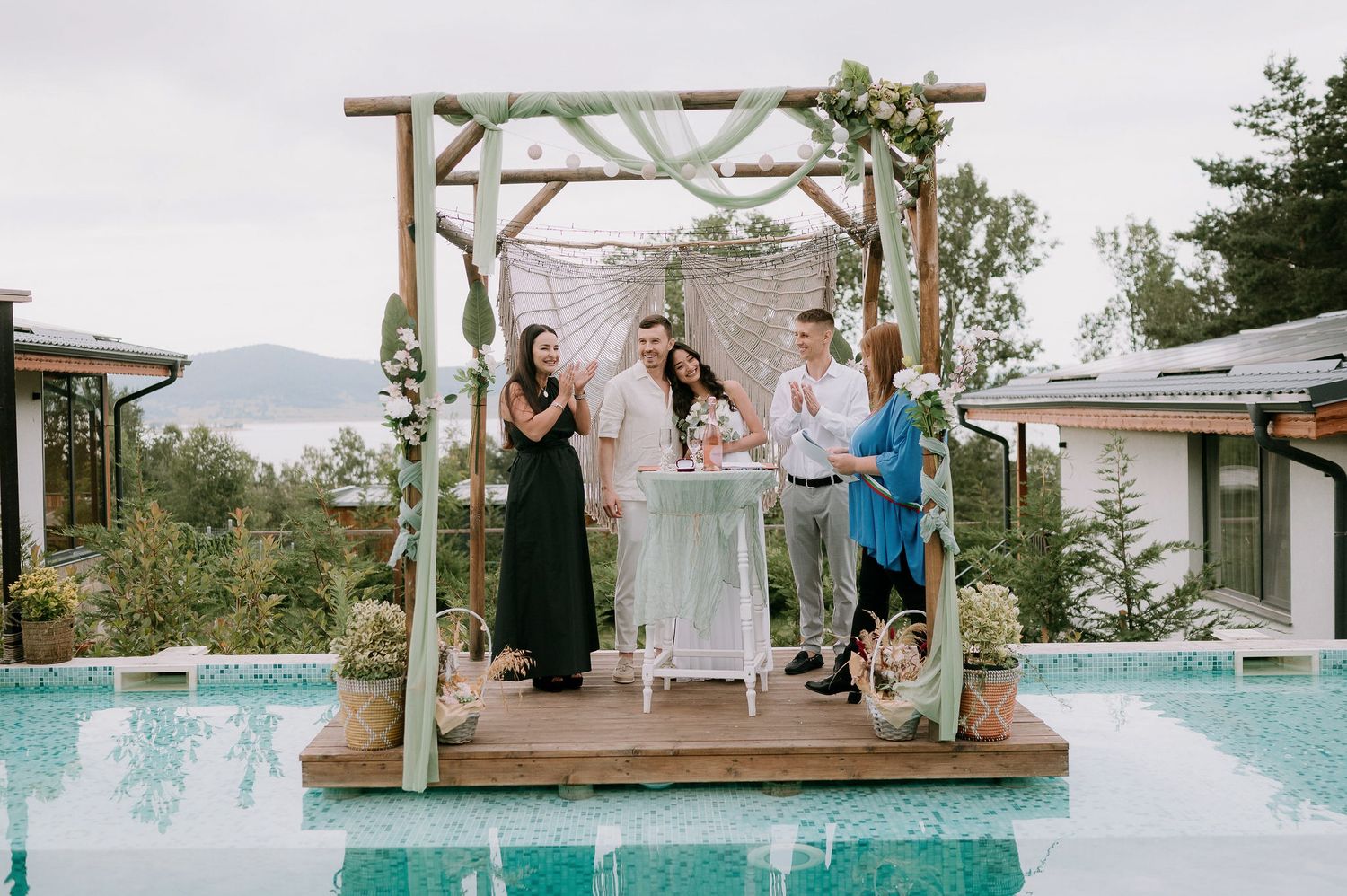 Group gathering for celebration under decorated pergola beside swimming pool.
