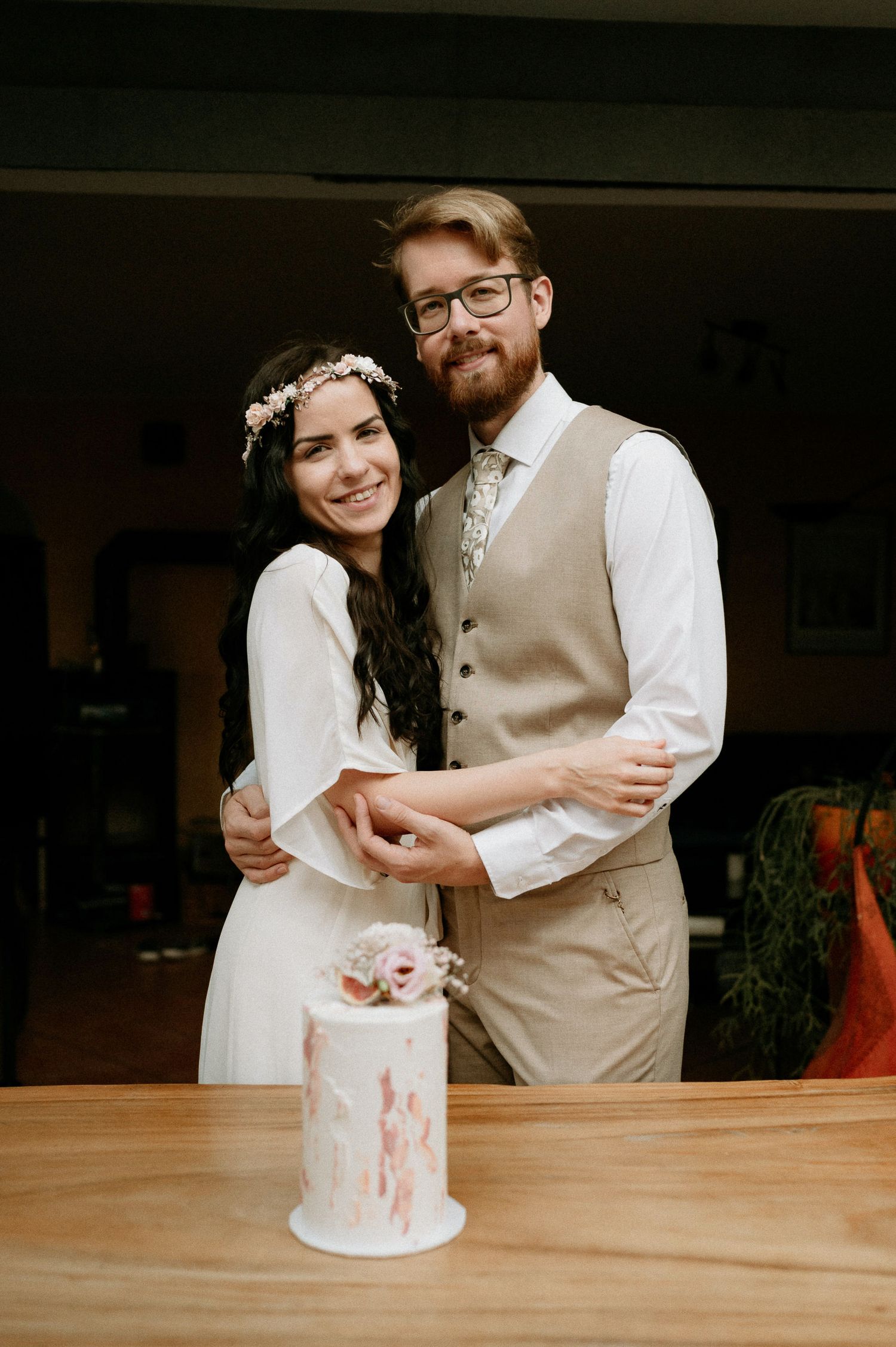 A couple stands behind their small wedding cake on a wooden table during an intimate celebration.