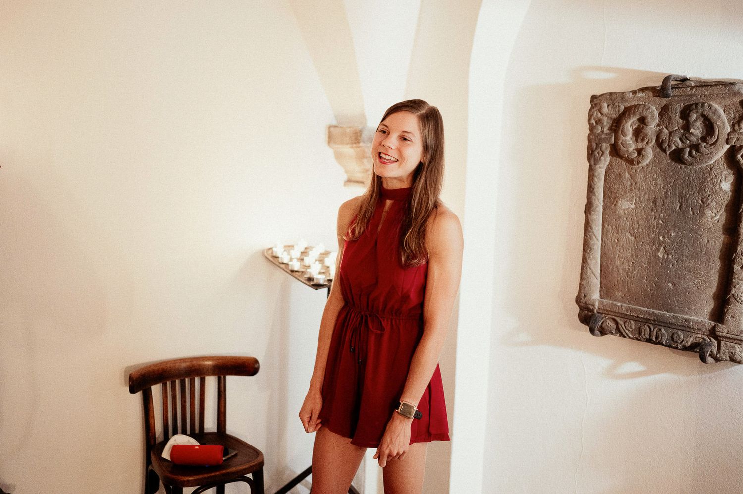 Person in red dress holding candle in arched white interior space.