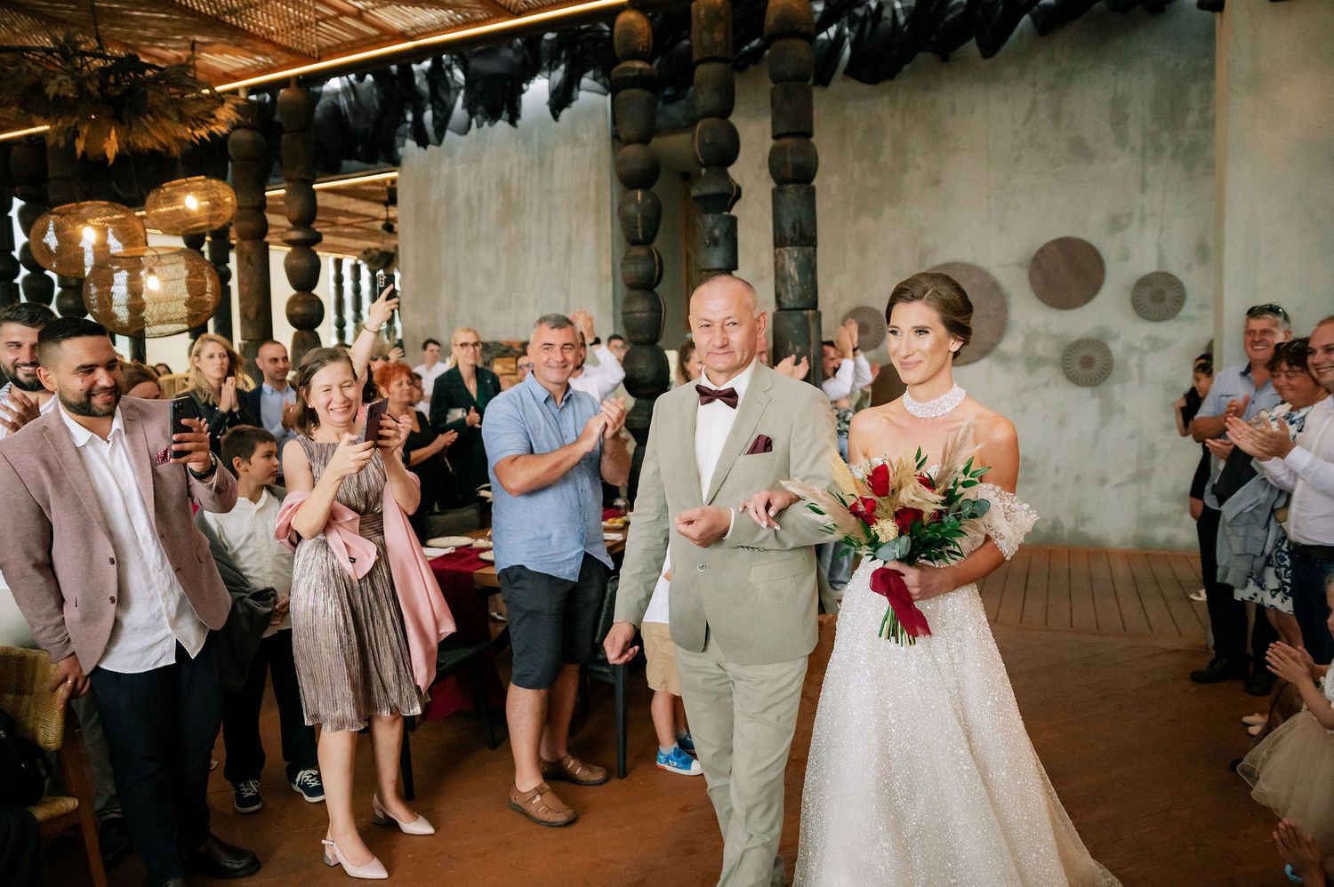 Wedding party and guests gathered in a rustic indoor venue with wooden ceiling beams.