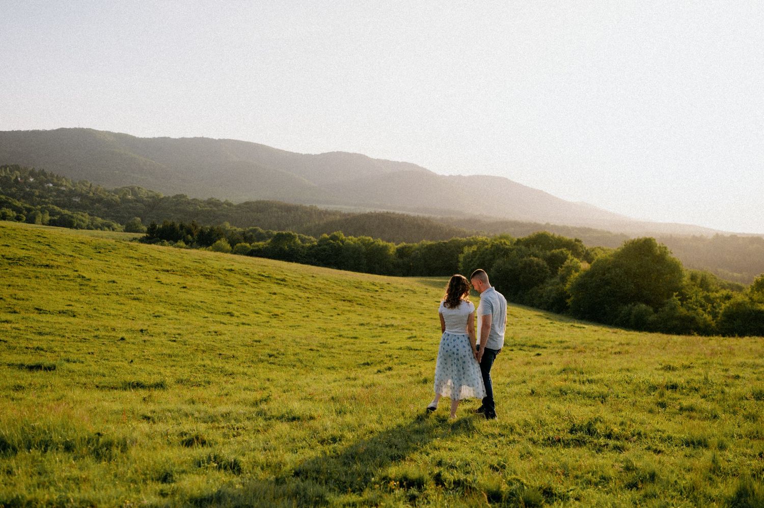 A couple shares a tender moment in a sunlit meadow with mountains in the distance.