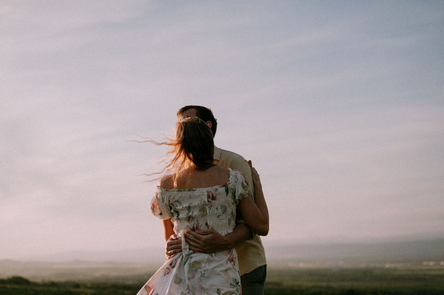 A couple embraces while standing in a scenic hilltop meadow at dusk.