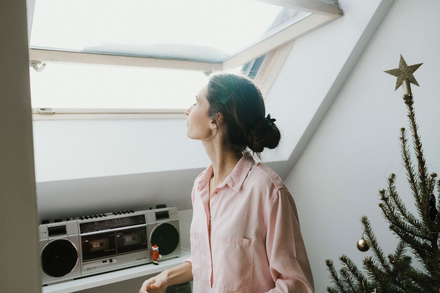 A person in a pink shirt gazes up at natural light coming through a skylight window, with a vintage boombox nearby.